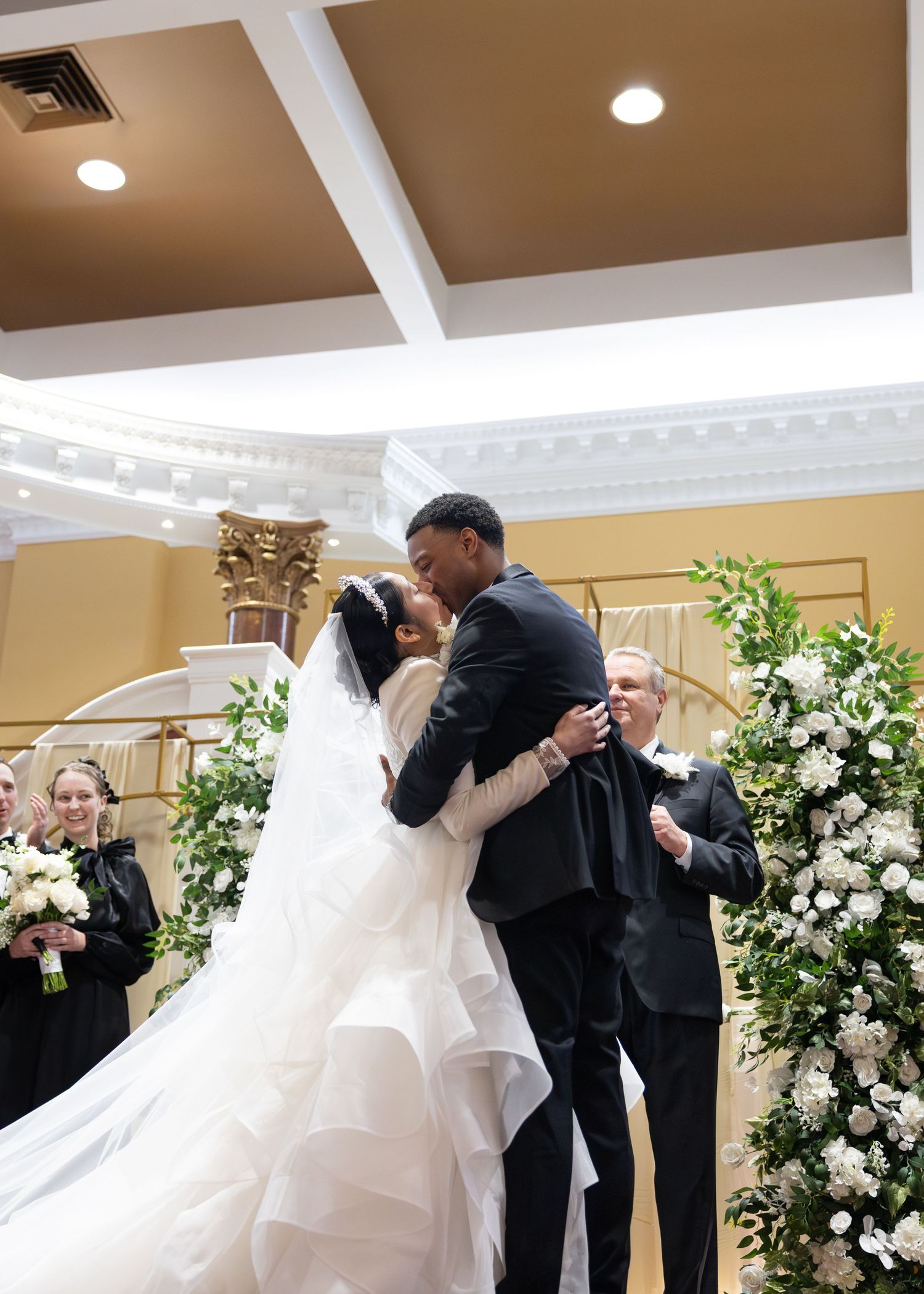 Bride and groom share first kiss at wedding altar adorned with lush white floral arch decorations.