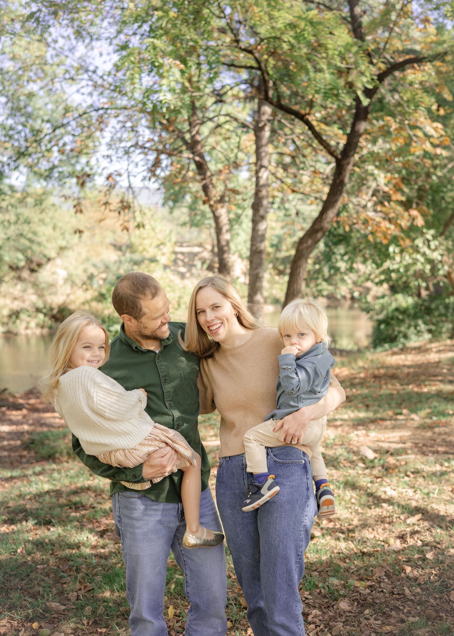 A happy family of four poses outdoors in autumn, parents holding two young children among trees with fall foliage. Roanoke, VA