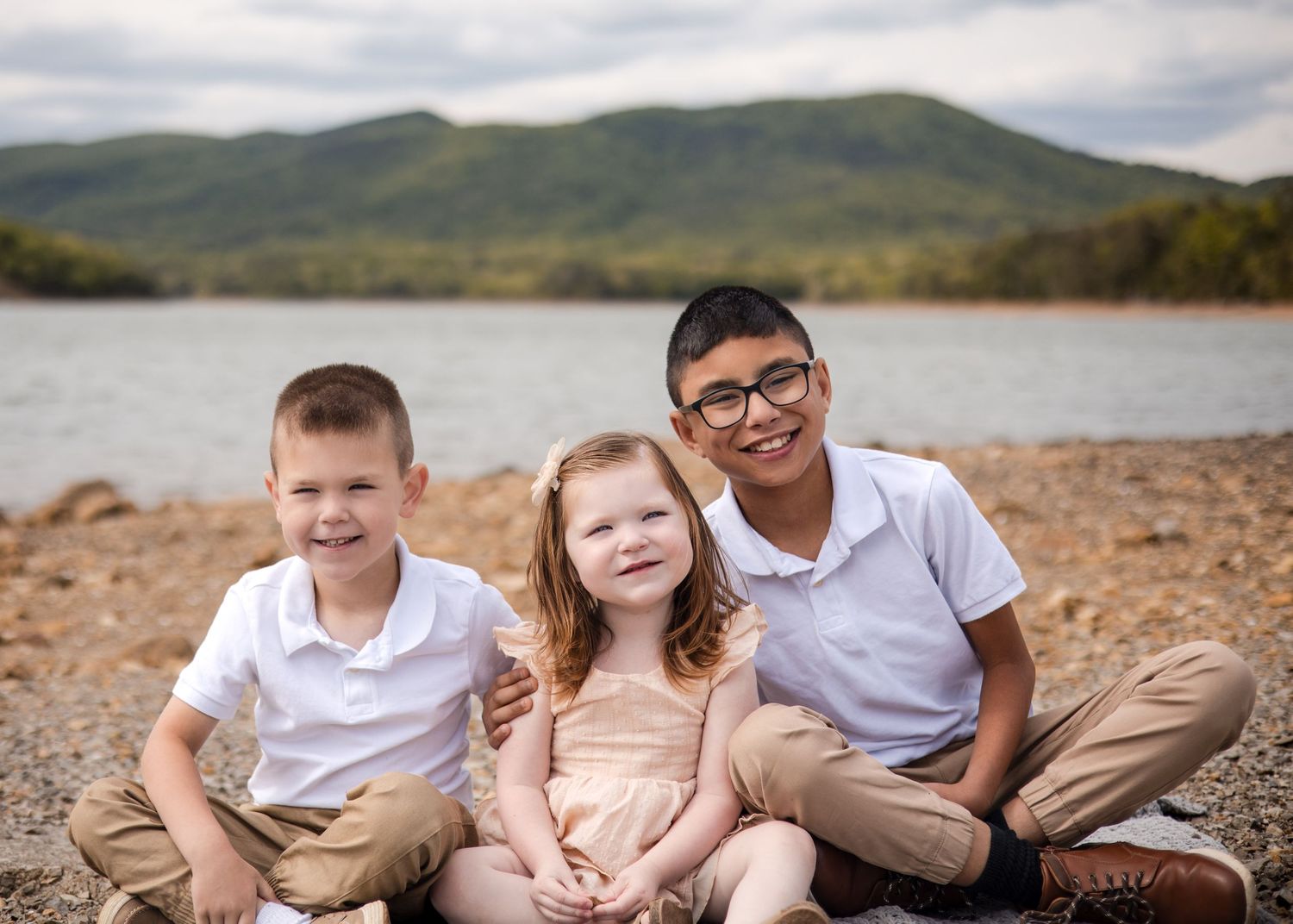 Three children smile together on a rocky lakeside beach with mountain views in a professional family portrait.