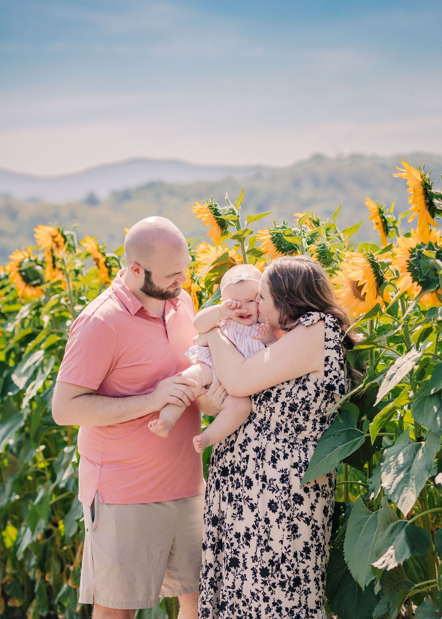 Family with baby posing in sunflower field with mountain backdrop on sunny day.