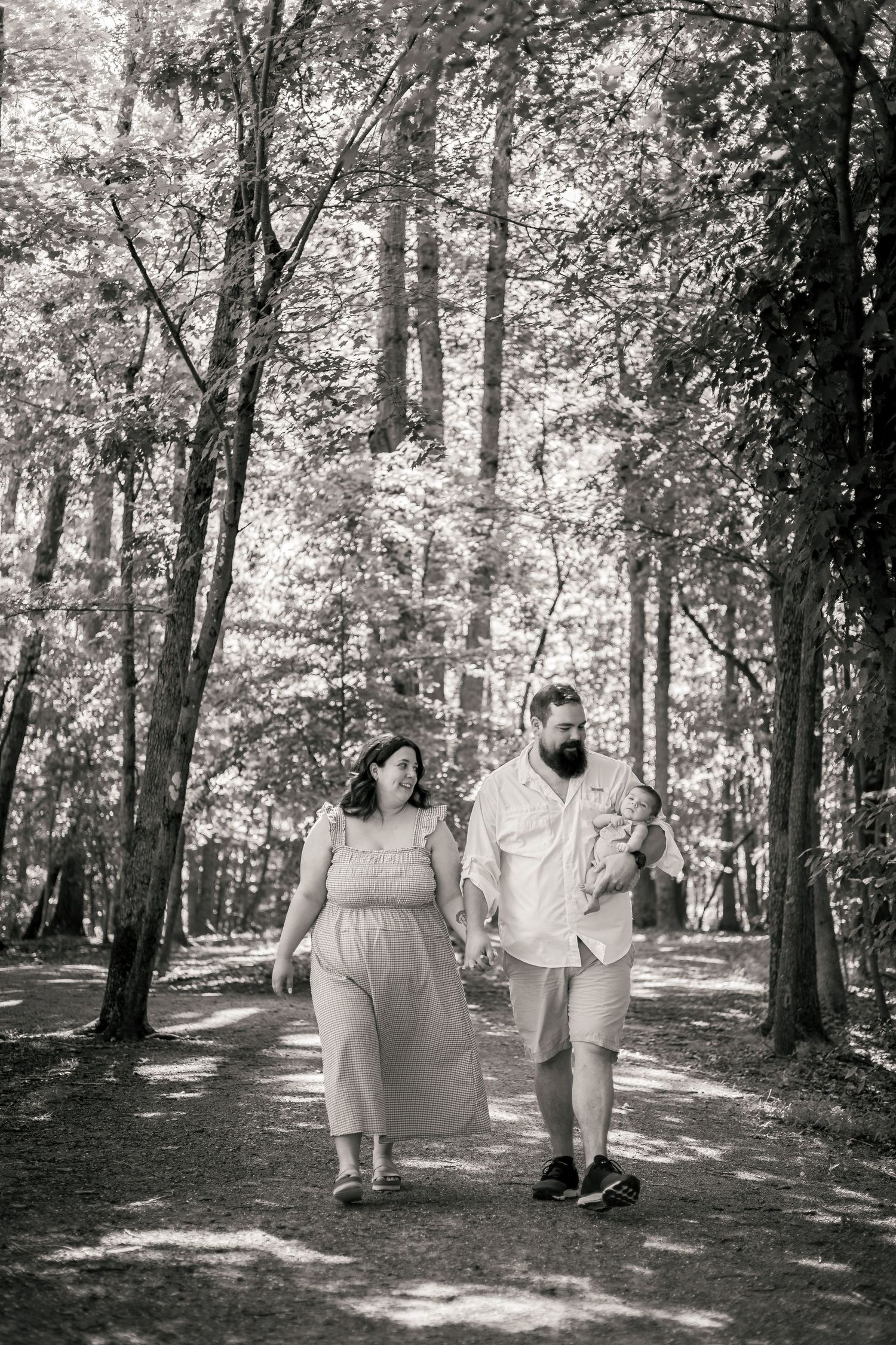 A couple walks hand-in-hand through a sunlit forest path, the man carrying a baby, in a black and white photo.