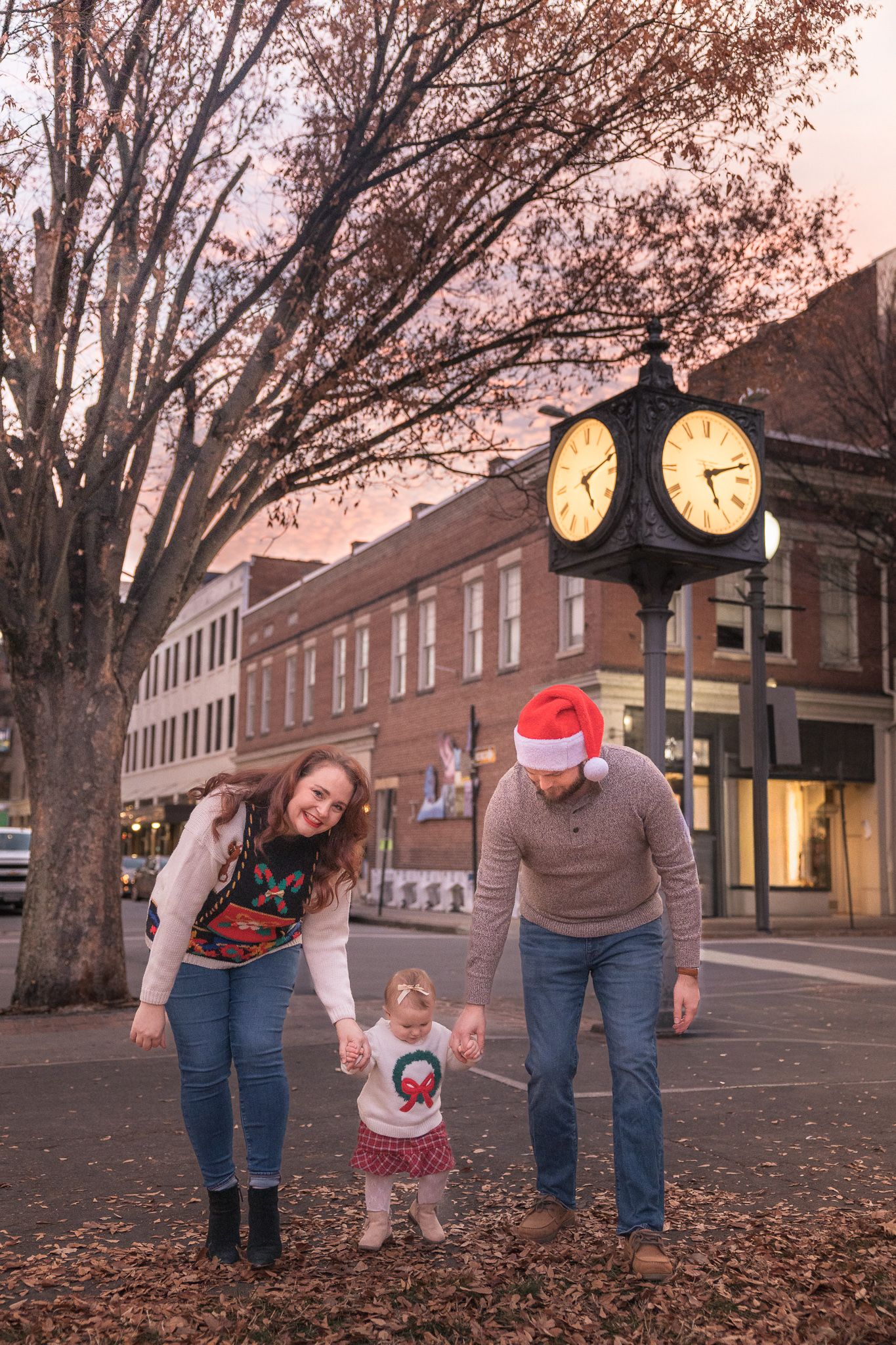 A family with a toddler celebrates Christmas outdoors near a vintage street clock at dusk in a festive town square.