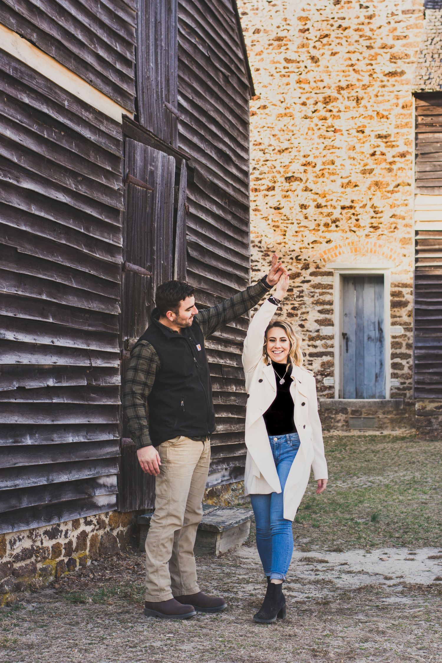Couple holding hands and dancing outside a rustic barn and stone building on a cool day.