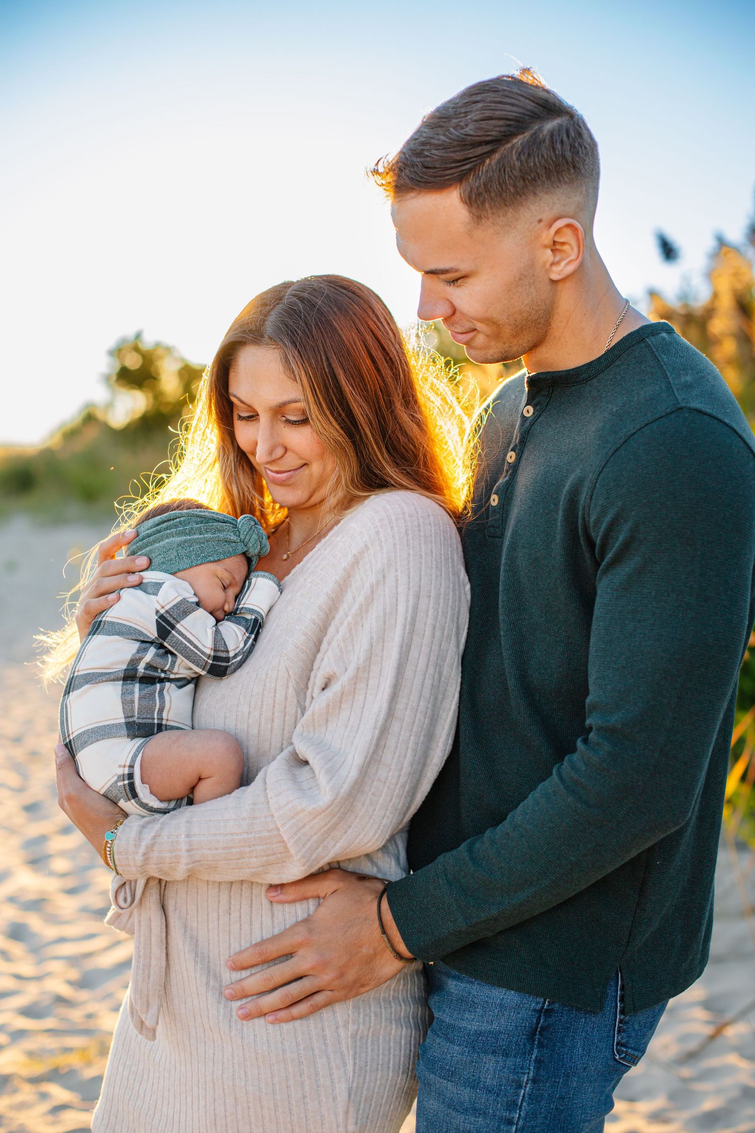 A couple stands on a sandy beach at golden hour, lovingly gazing at their newborn baby.