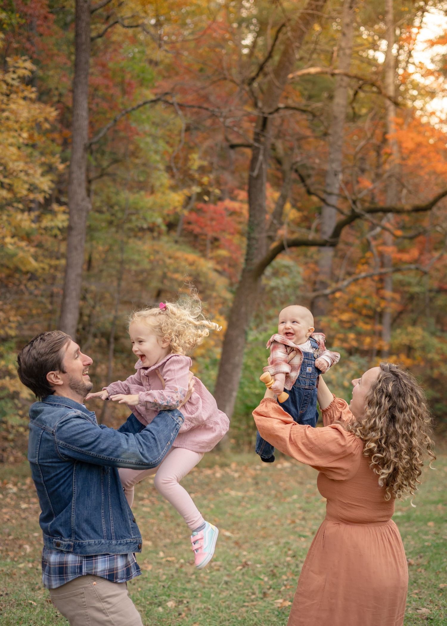 Family playing in autumn park, parents lifting two young children amid colorful fall foliage.