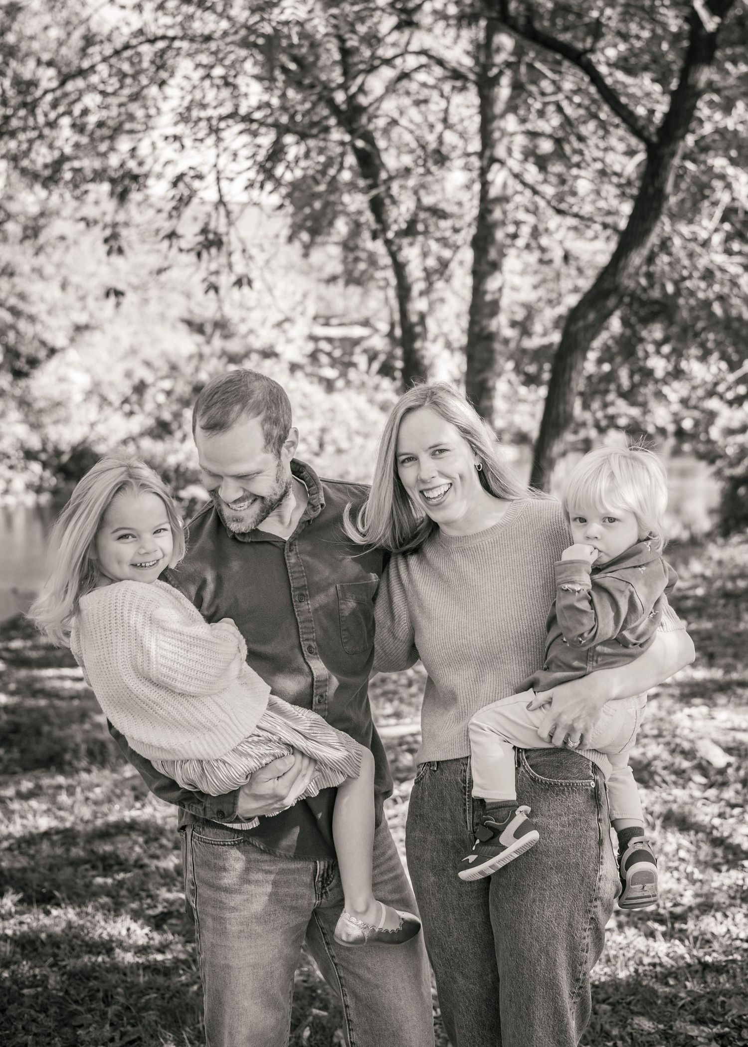 Black and white family portrait of parents holding two young children outdoors among trees.
