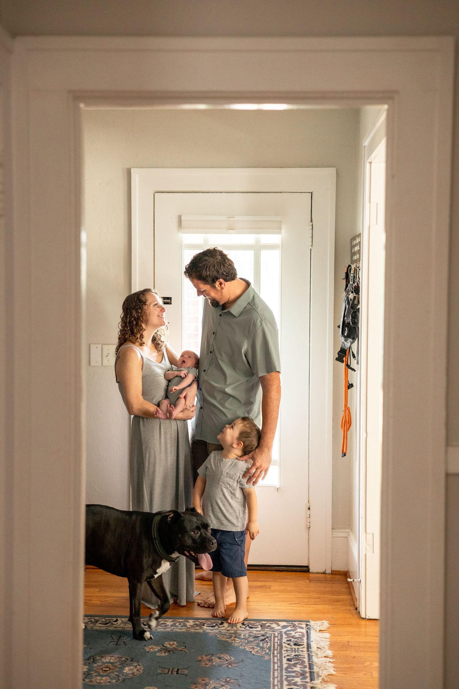 A young family with a newborn, toddler, and black dog stand together in a bright home hallway.