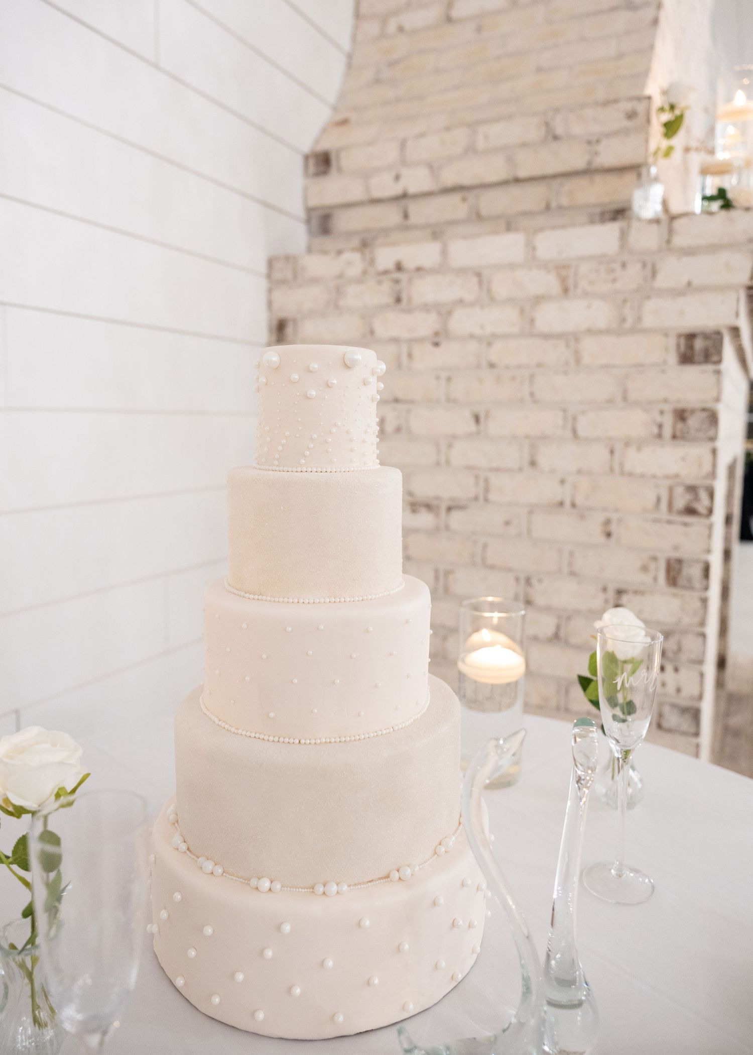 Elegant five-tier all-white wedding cake with pearl details on a table, set against a rustic white brick wall backdrop.