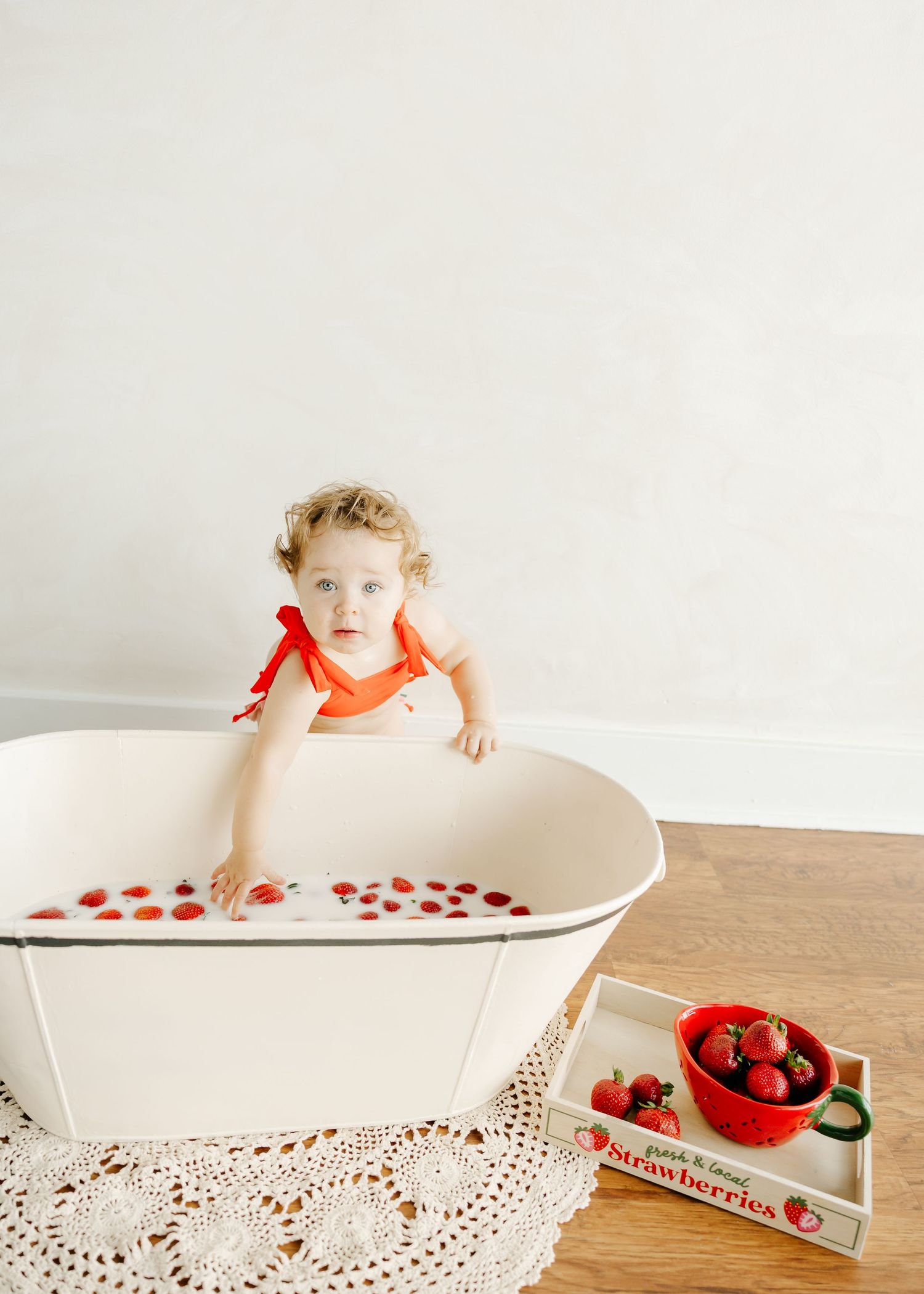 Toddler girl in red swimsuit sitting in white enamel tub with strawberries, next to a tray of fresh strawberries.