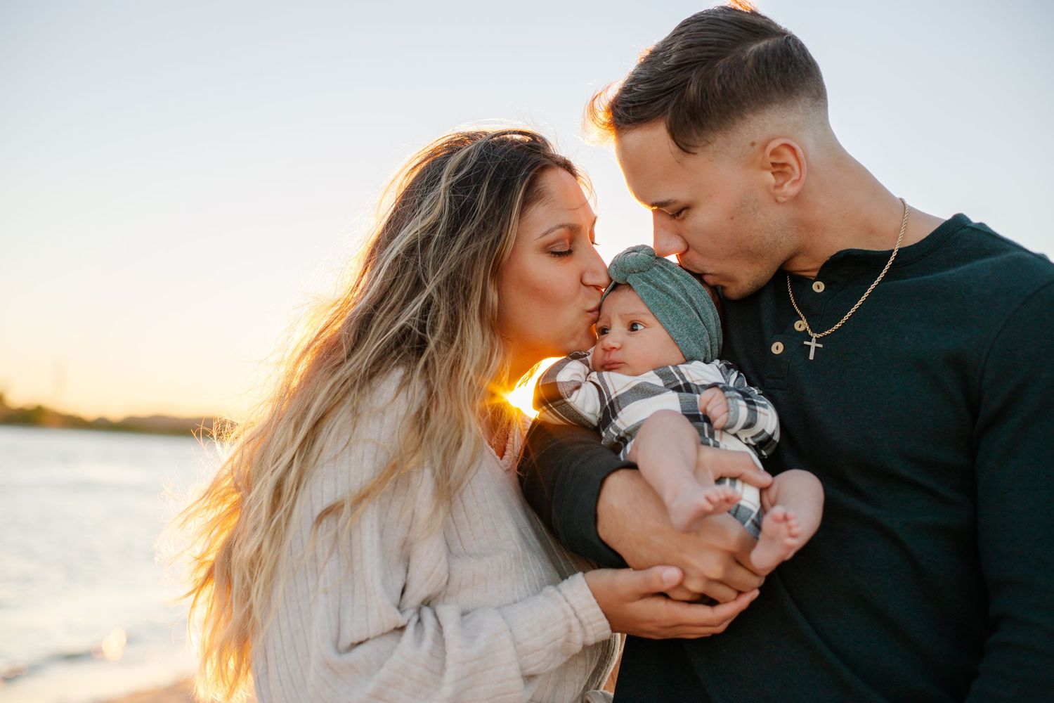 Couple kissing newborn baby on beach at golden sunset, family portrait outdoors by the water.
