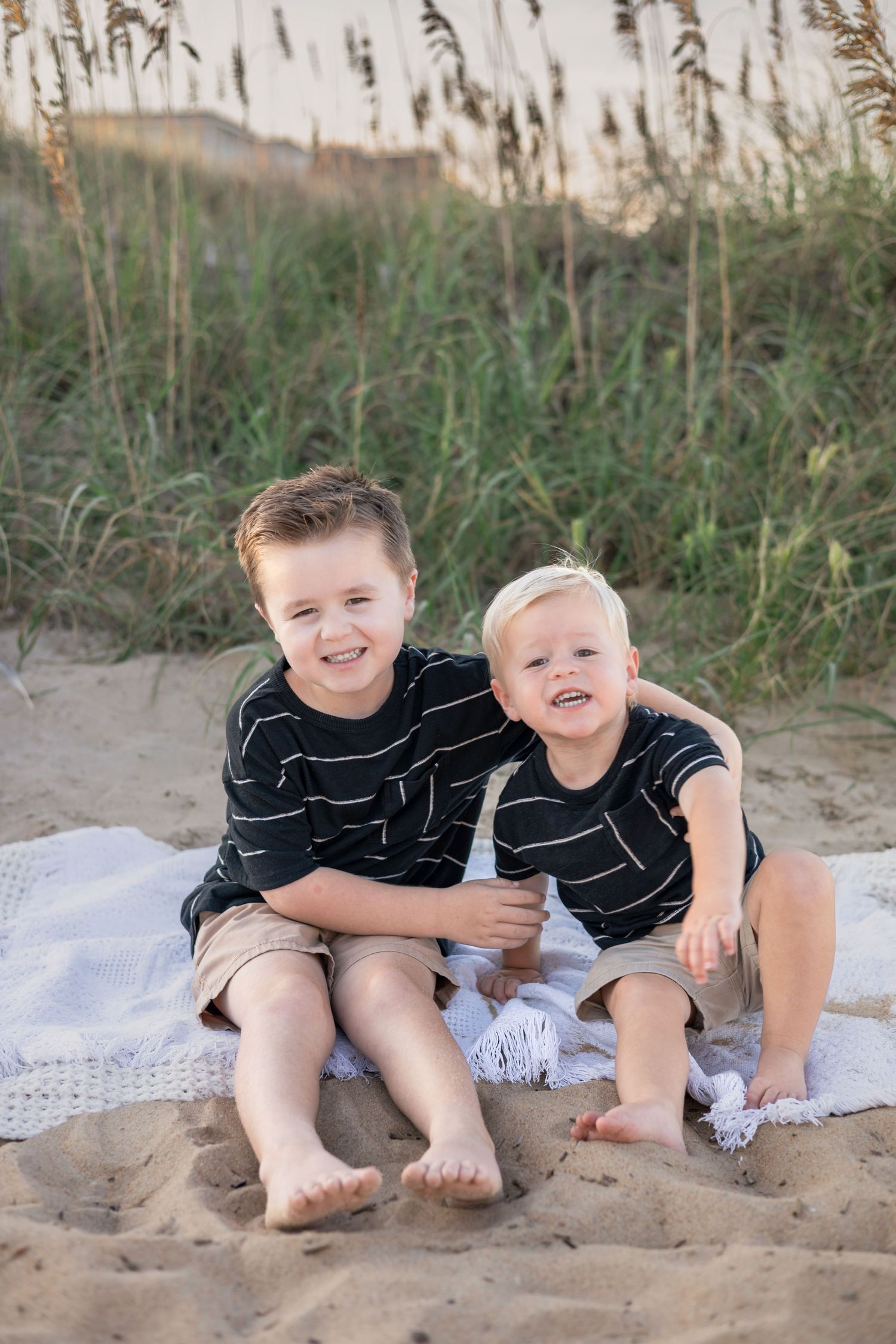 Two young brothers in matching black striped shirts sit together on a sandy beach blanket with sea oats behind them.
