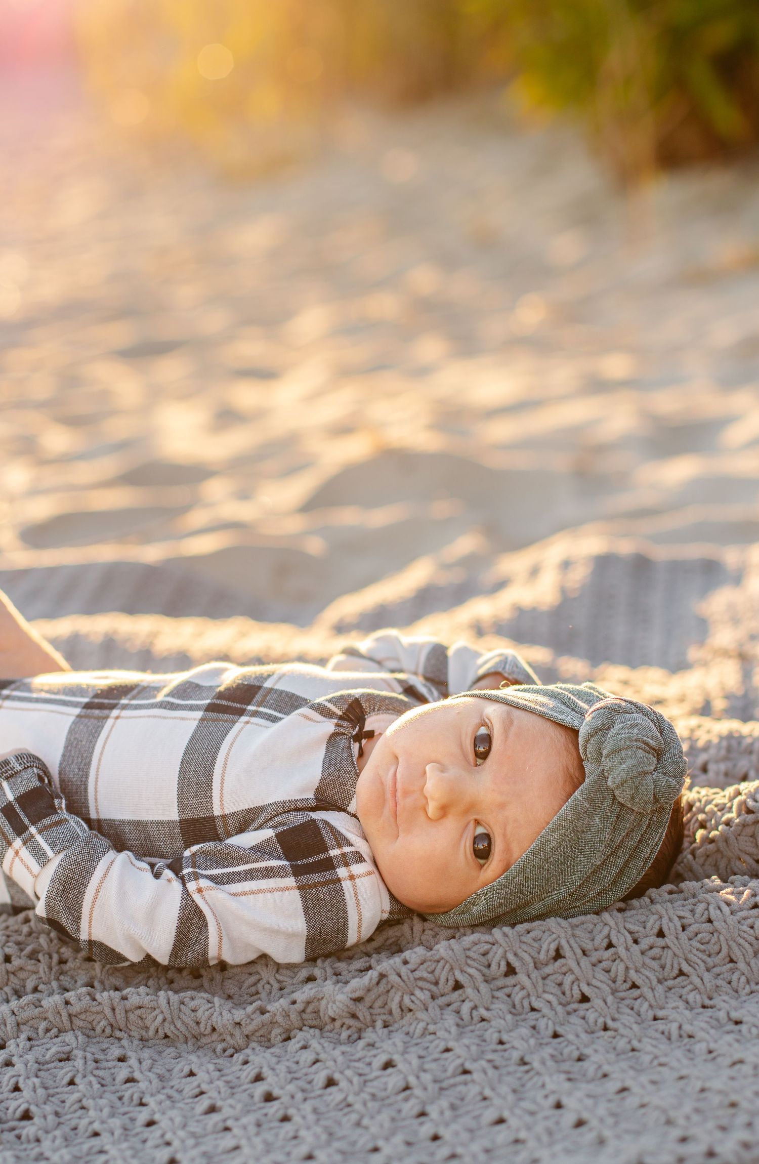 A baby in a plaid outfit and knit hat lies on a crochet blanket on sandy beach in warm golden light.