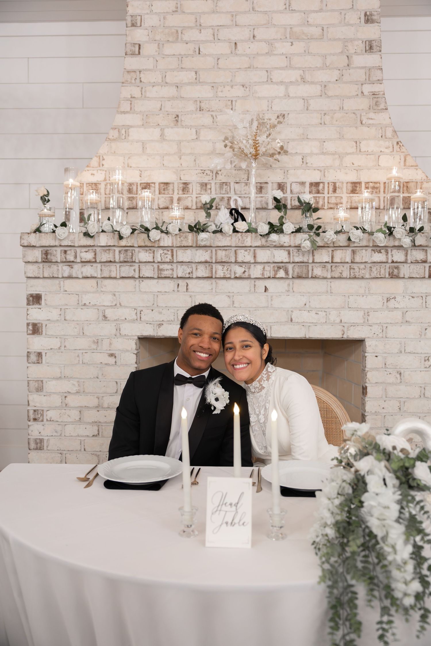 Newlywed couple smiling at head table with white brick fireplace backdrop, candles, and floral arrangements at wedding reception.