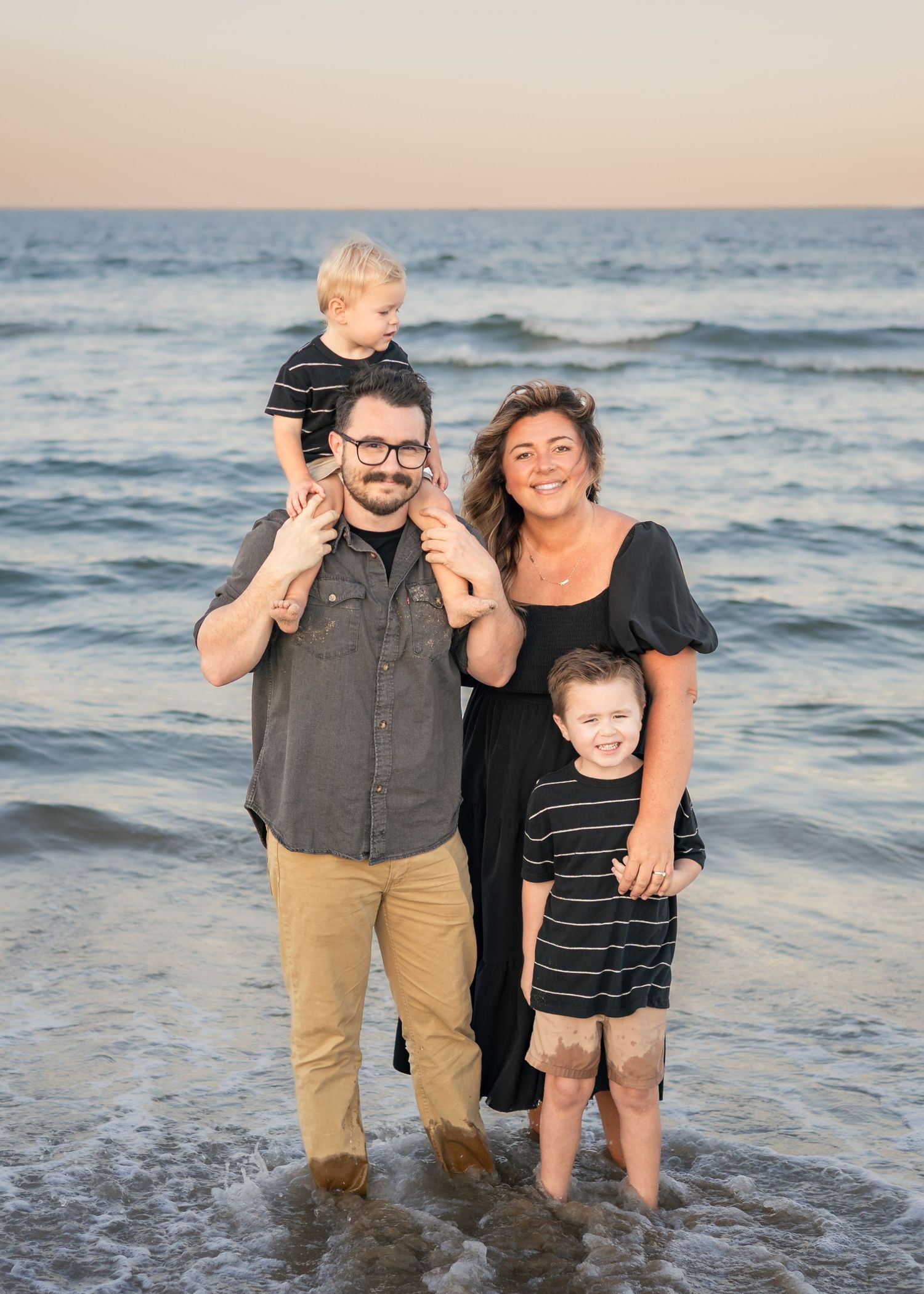 Family of four smiling on a beach at sunset, father holding toddler on shoulders, mother beside older child.