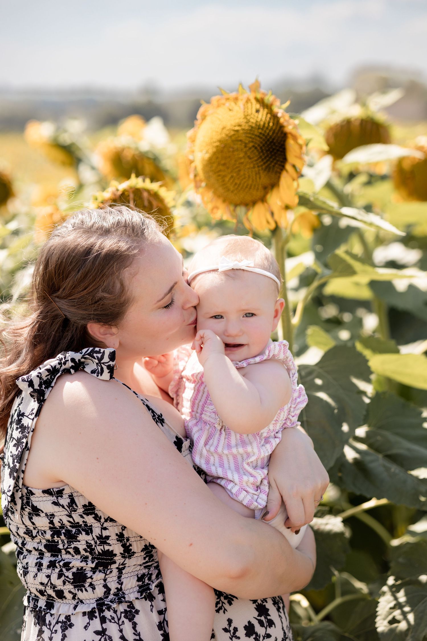 Mother kissing baby girl in sunflower field on sunny day.