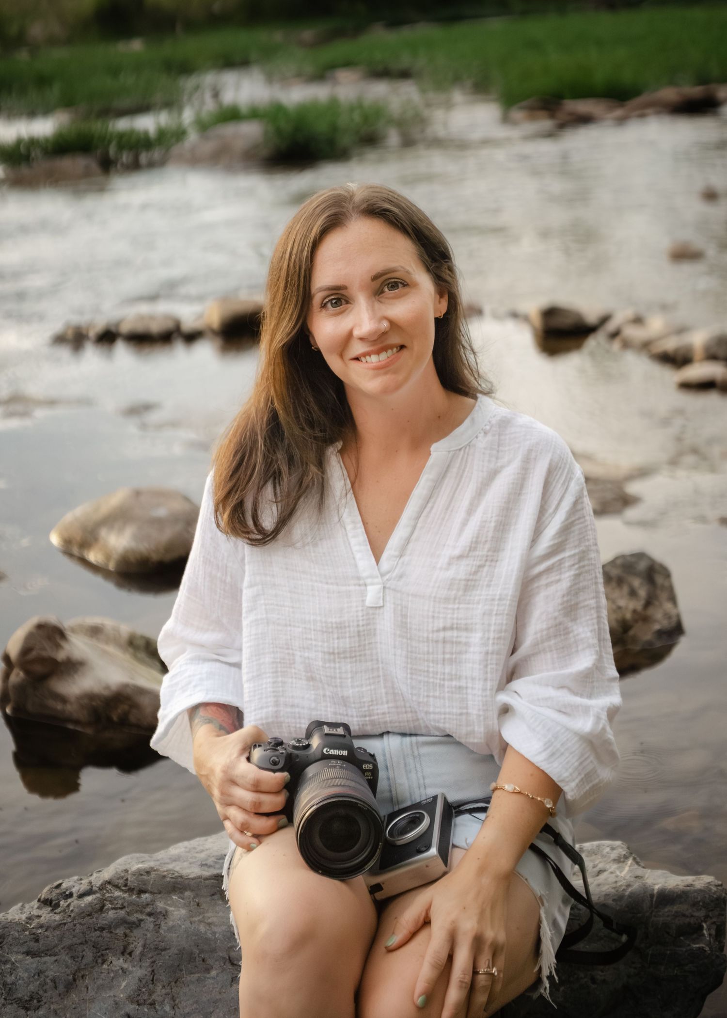 Woman photographer smiling, holding a camera, sitting on rocks by a serene river in a white linen shirt.