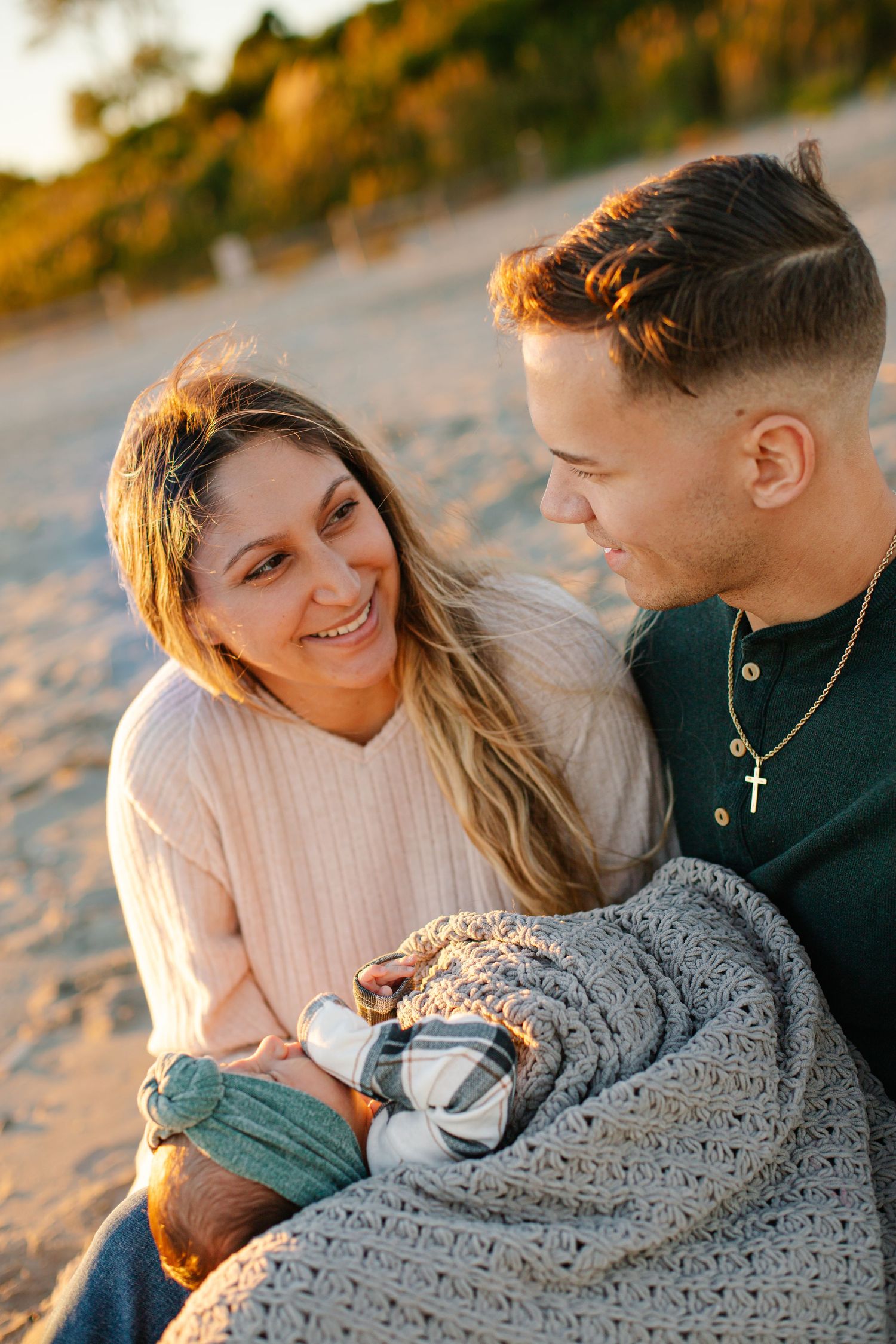 A smiling couple wrapped in a cozy blanket sits on a sandy beach at golden hour, sharing a warm and intimate moment.