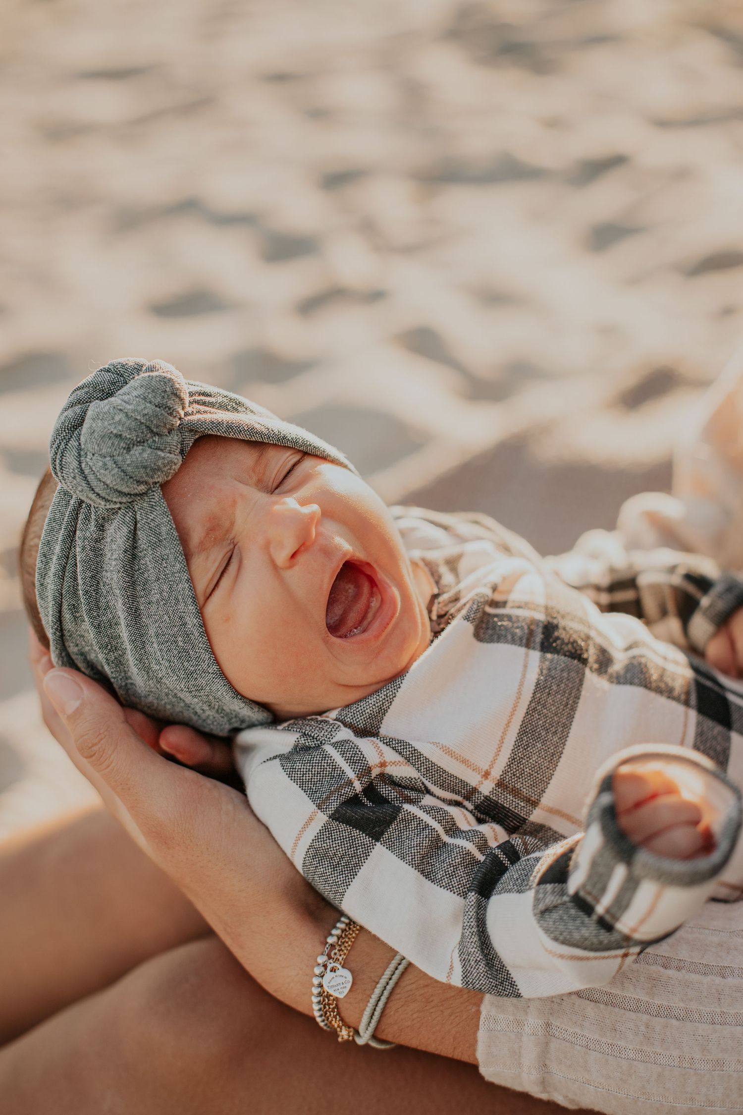 A newborn baby yawning while being cradled, wearing a plaid outfit and green turban hat on a sandy beach.