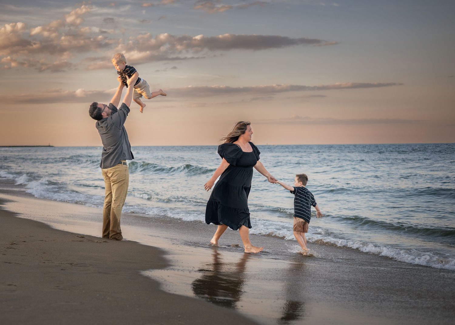 Family enjoying a beach sunset, father tossing baby in air, mother and young son playing near ocean waves.