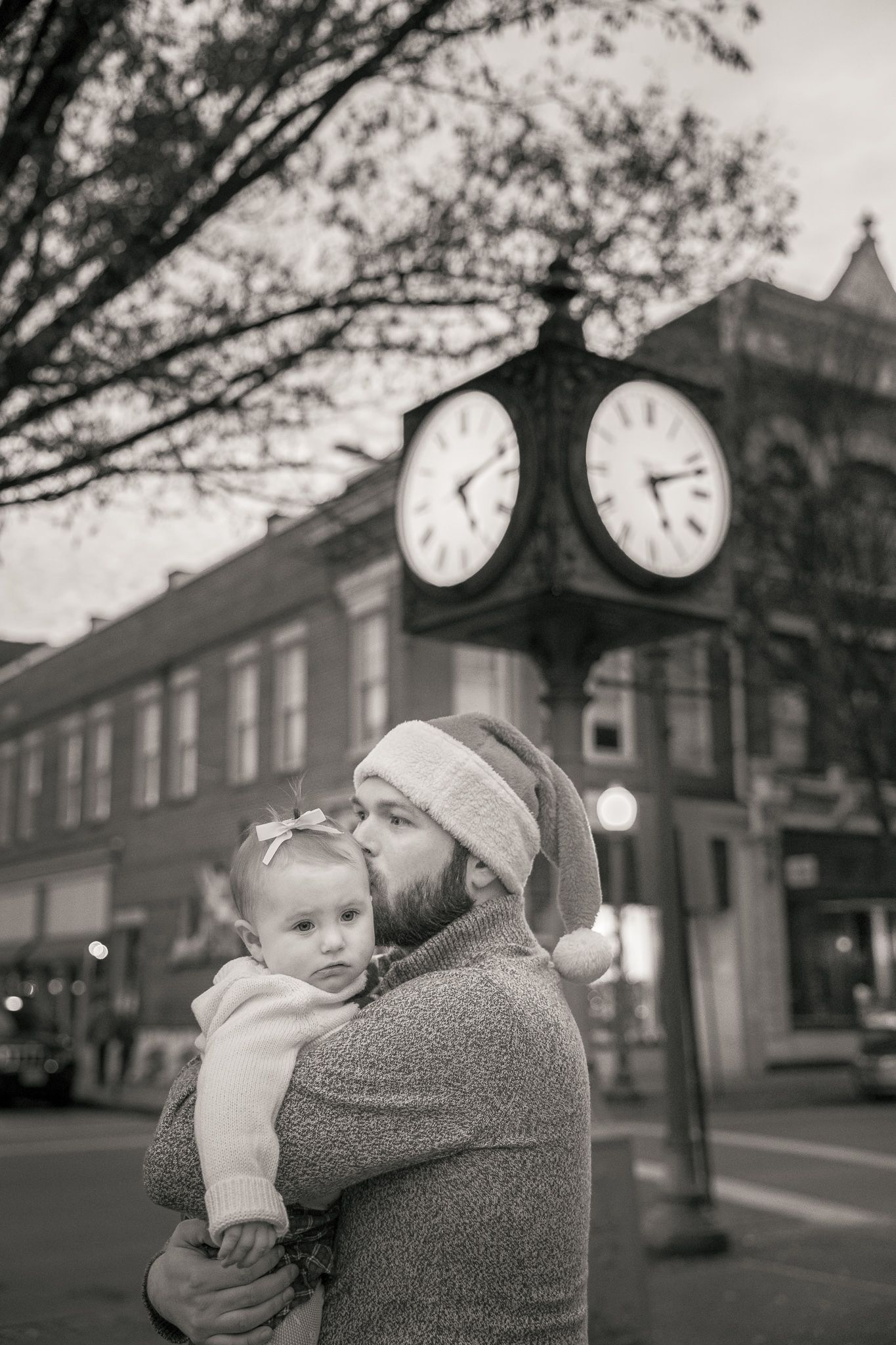 Black and white photo of a bearded man in Santa hat kissing baby girl near vintage street clock at Christmas.