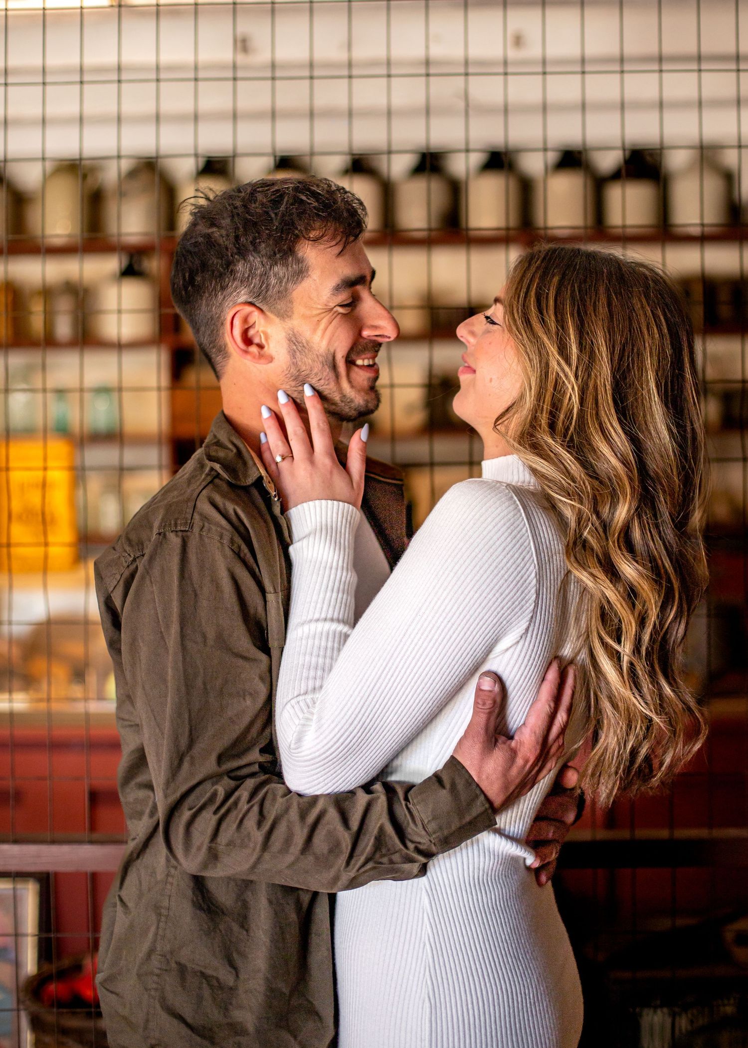 A couple embraces and smiles at each other in a cozy indoor setting, the woman in white and man in olive jacket.