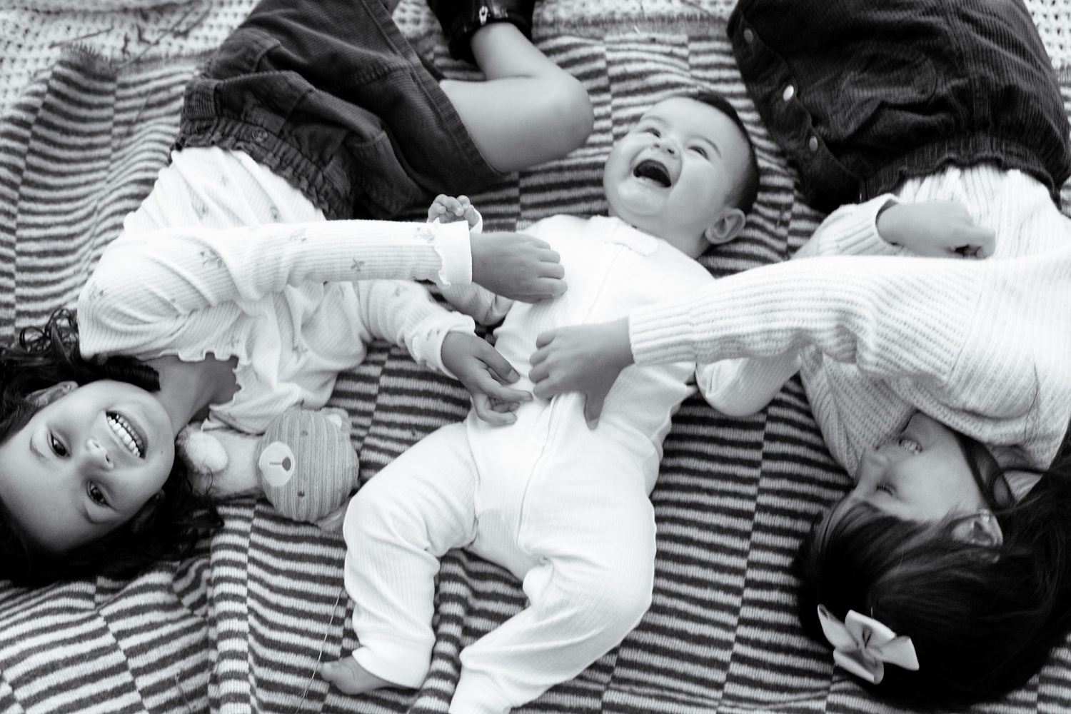 Black and white photo of three young children lying on a striped blanket, with a laughing baby in the center.