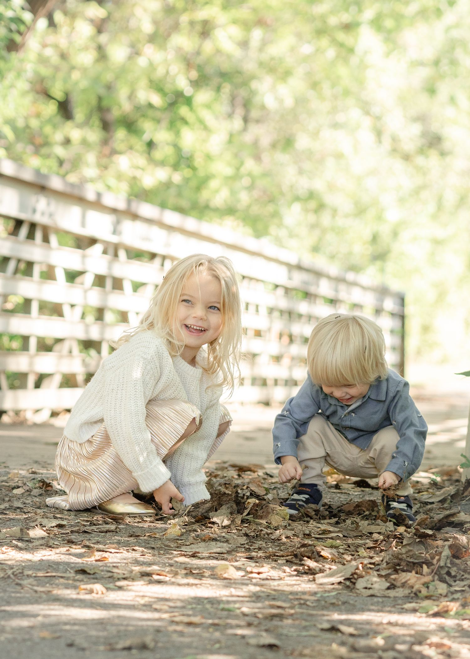 Two young blonde children playing with leaves on a dirt path near a white wooden fence outdoors.