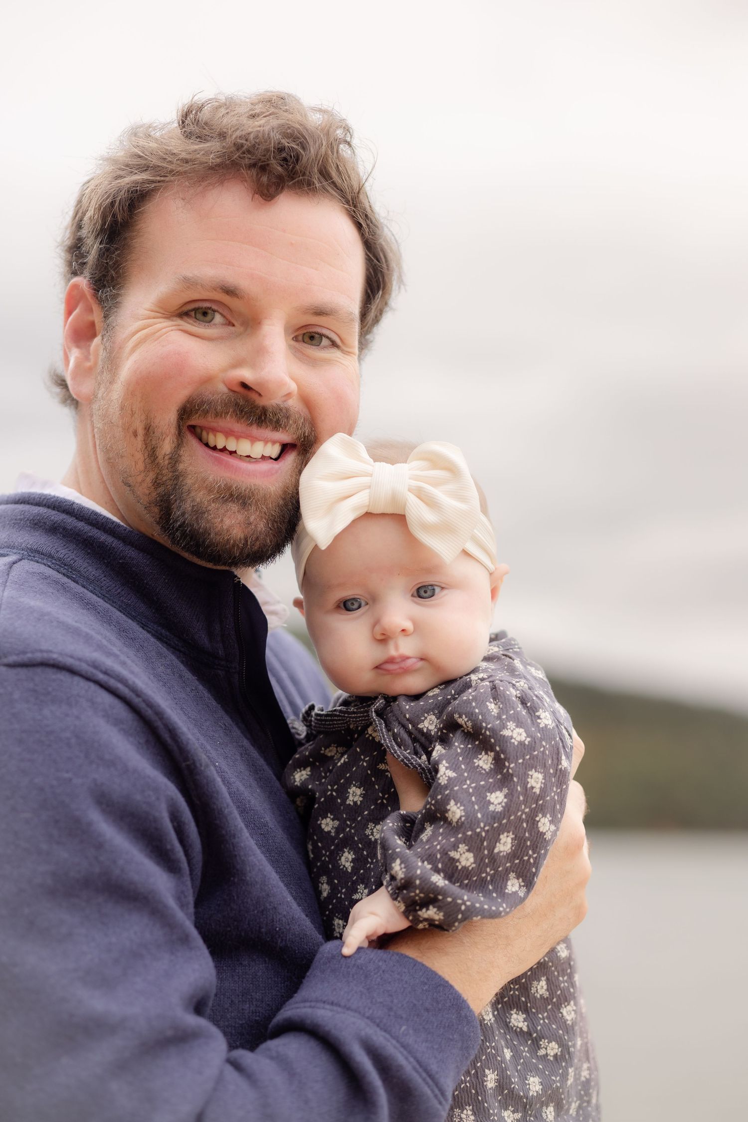 Smiling bearded father holding his baby girl in a floral dress and bow headband outdoors.