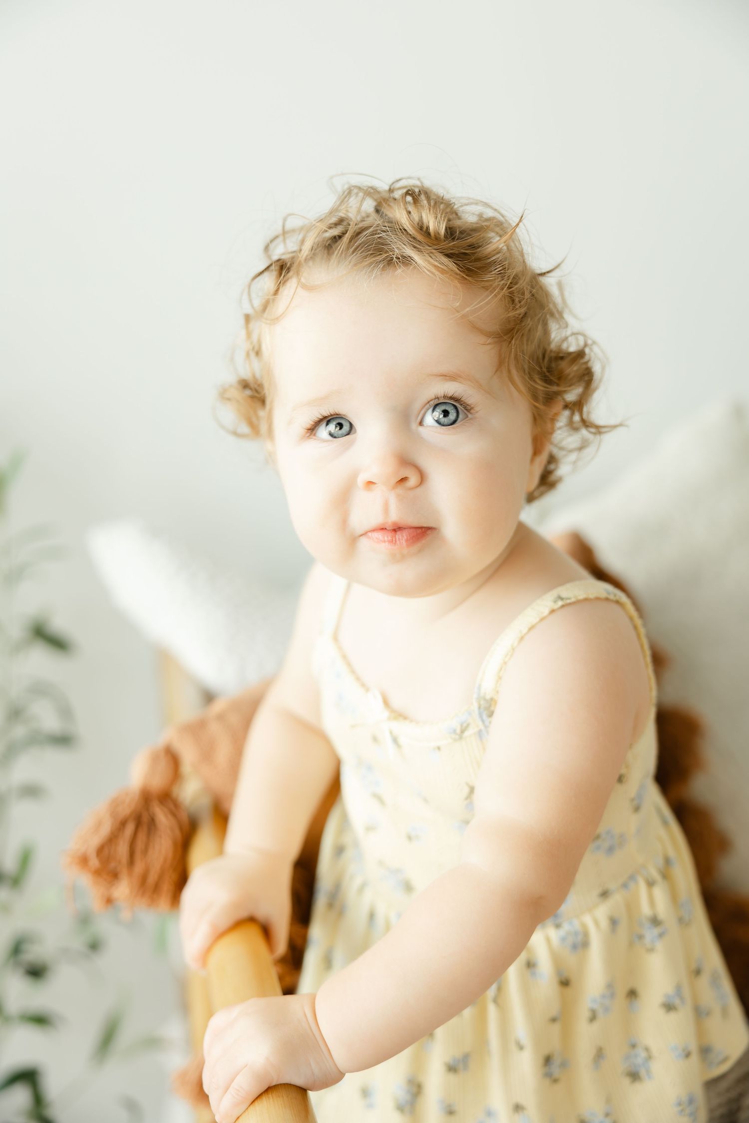 Cute baby girl with curly hair and blue eyes in a floral yellow dress holding a wooden rocking horse toy.