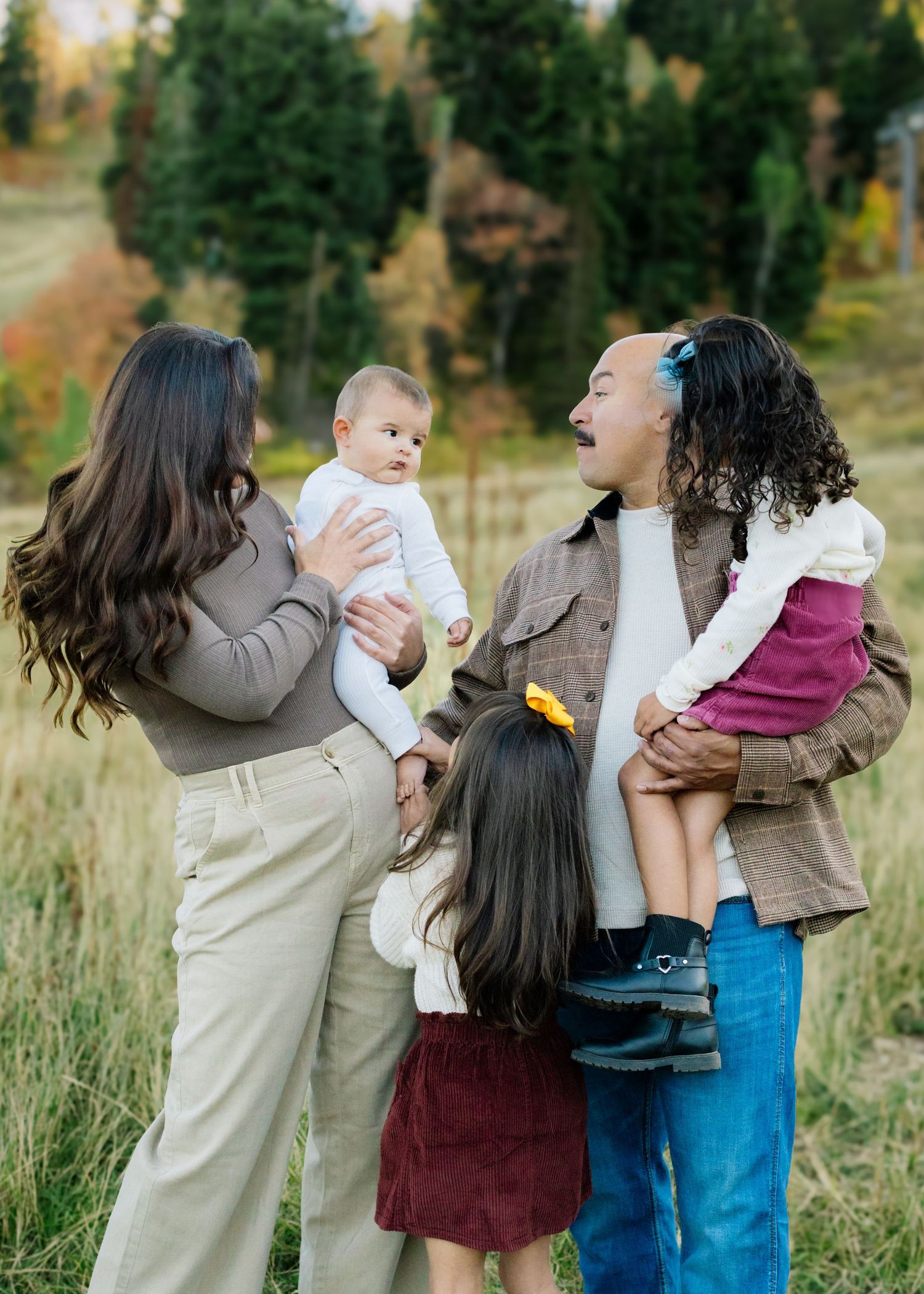 Family of four with two young children posing outdoors in a meadow with autumn trees in the background.