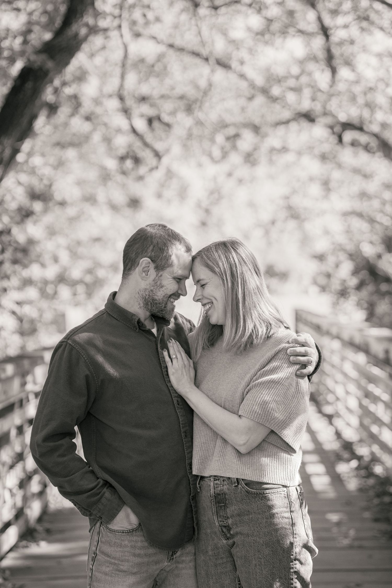 A couple shares a romantic moment on a bridge surrounded by autumn trees in a black and white photo.
