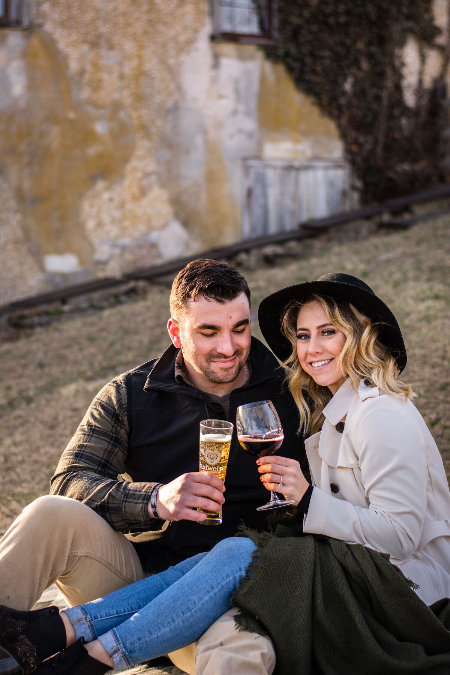 Couple sitting outdoors on grass, clinking wine glasses, smiling, wearing casual fall attire near a rustic stone building.