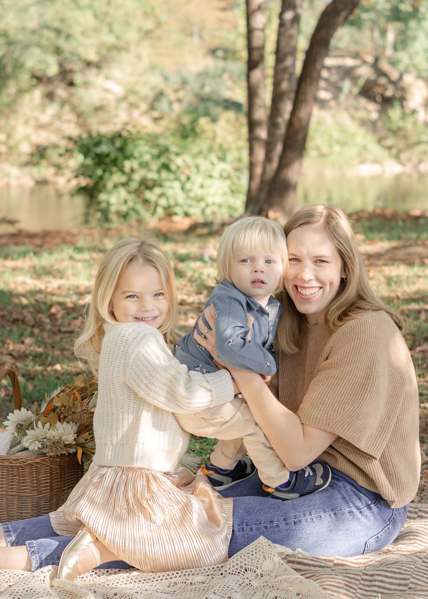 A smiling mother holds her toddler son while sitting outdoors with her blonde daughter during a fall family photo session.