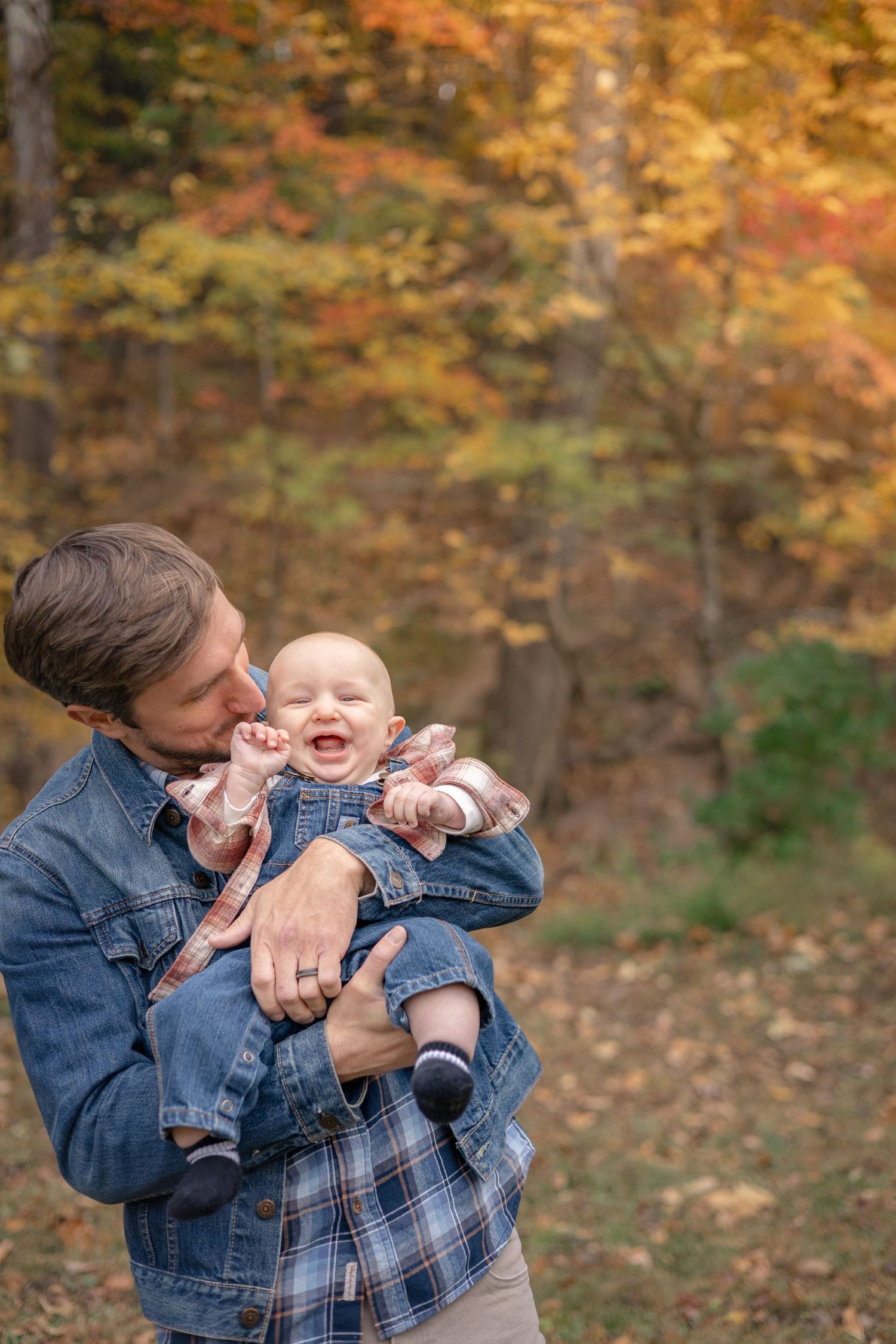 Father in denim jacket kissing laughing baby outdoors among colorful autumn foliage.