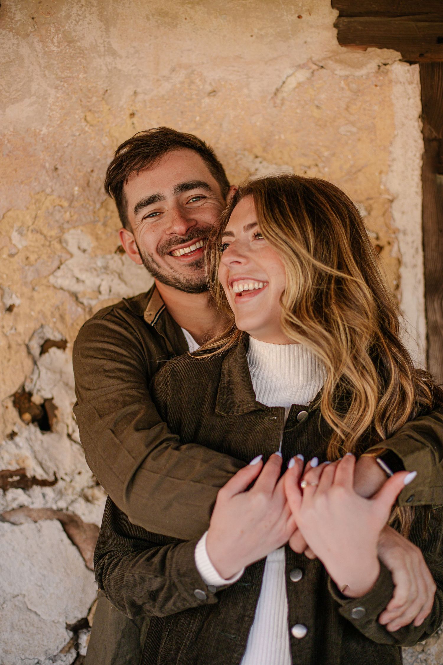 Happy couple embracing and laughing together against a rustic stone wall backdrop.