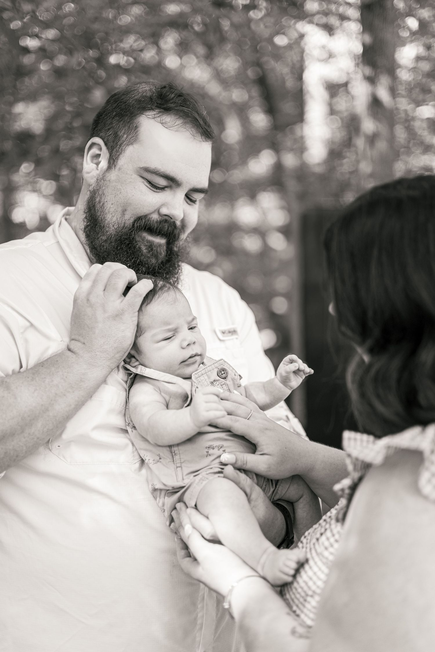 Black and white photo of a bearded father holding a newborn baby outdoors, with a woman reaching out to touch the infant.