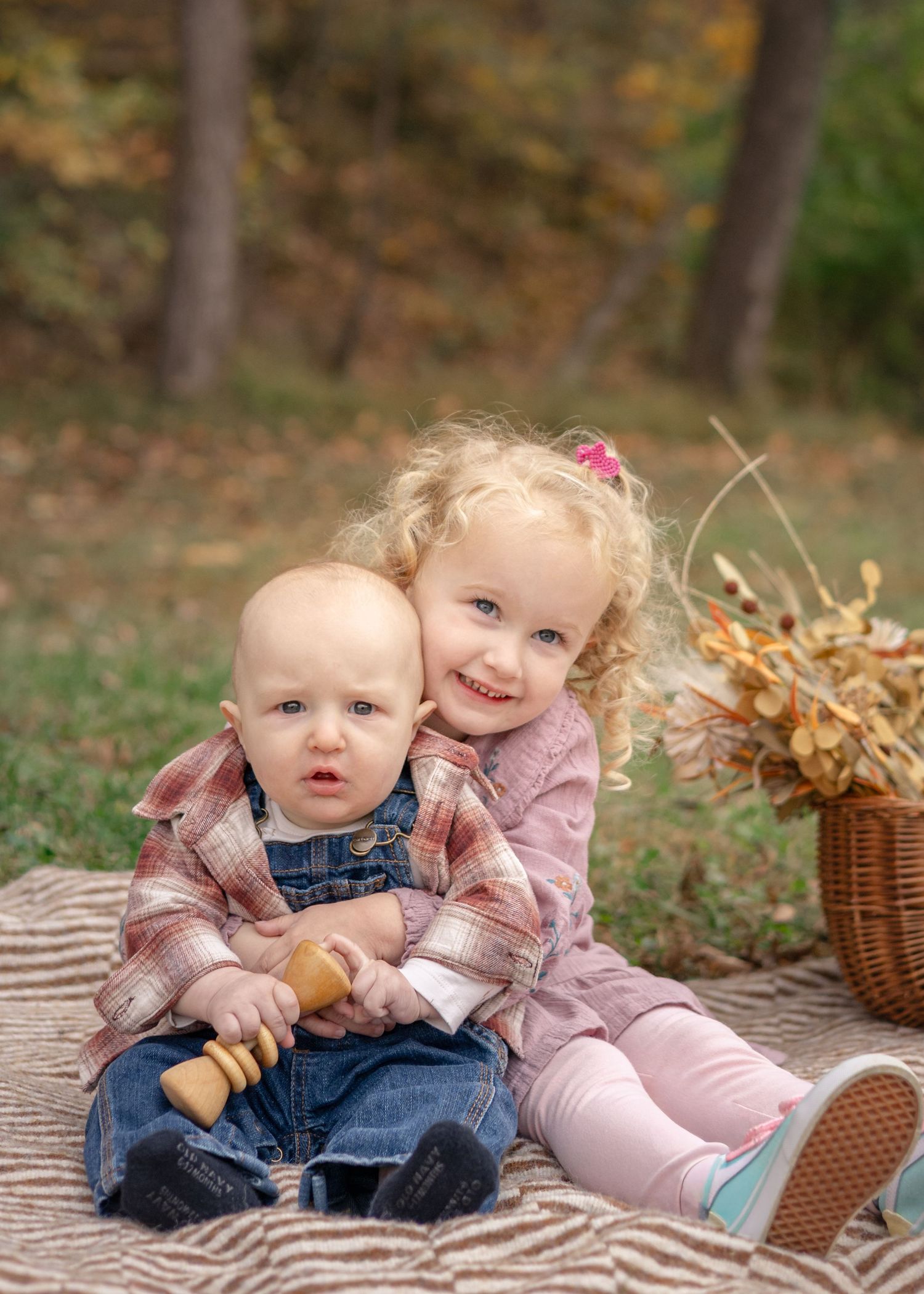Two young siblings sit together on a blanket outdoors in autumn, the older blonde girl hugging her baby brother.