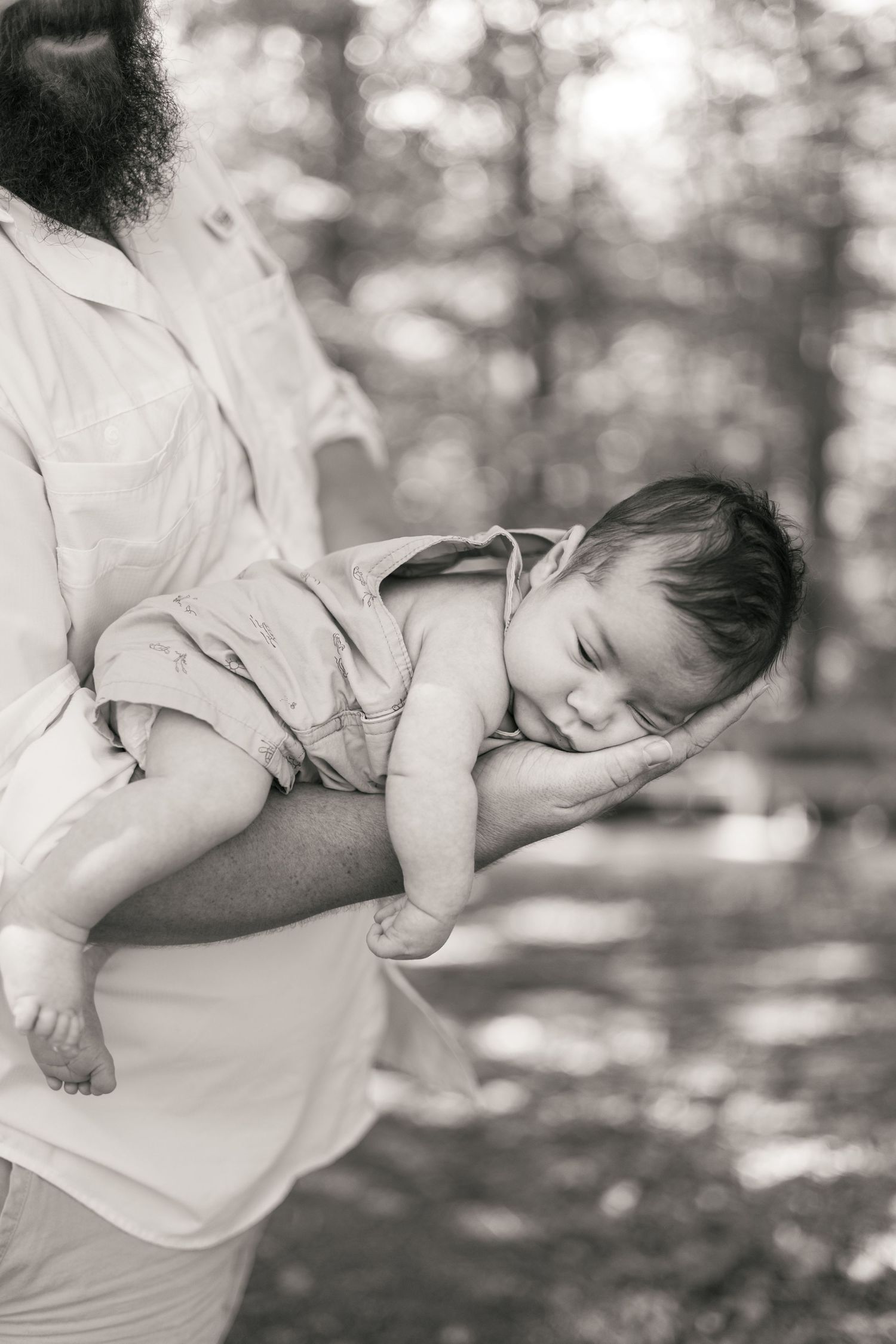 Black and white photo of a bearded father cradling a sleeping newborn baby outdoors in a sunlit forest.