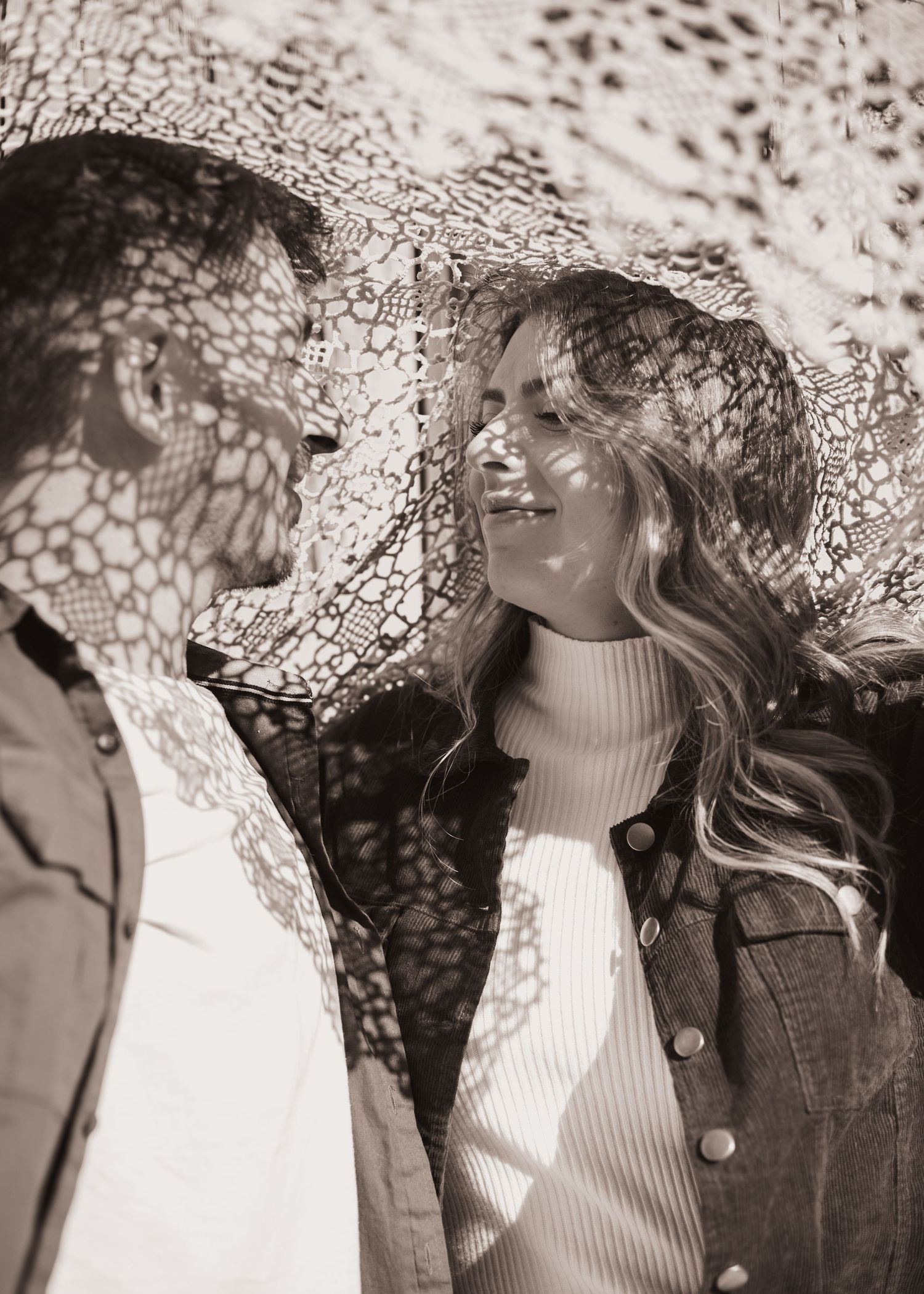 Sepia-toned couple smiling at each other under lace fabric casting delicate shadows in romantic outdoor portrait.
