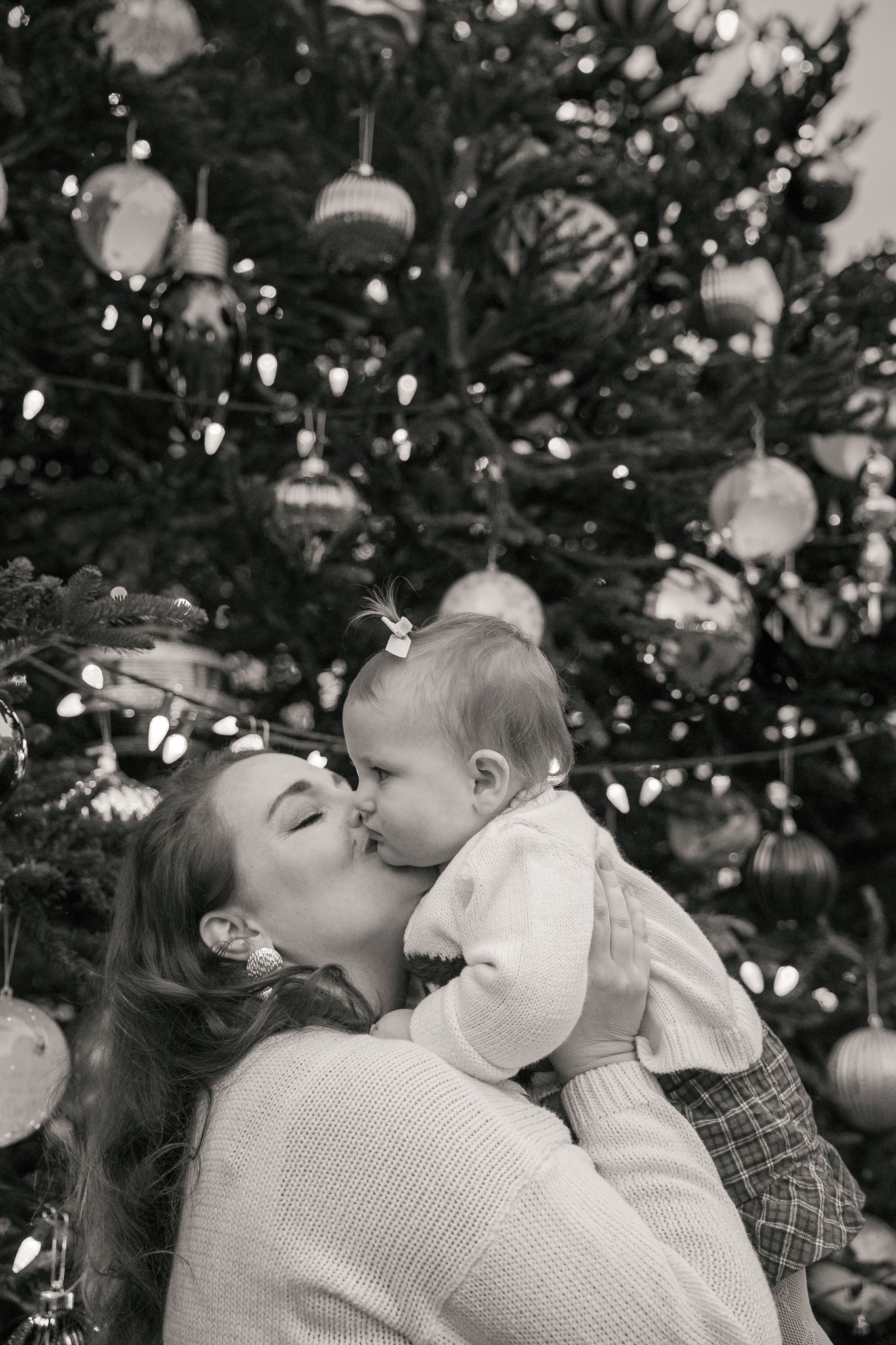 Black and white photo of a mother kissing her baby in front of a decorated Christmas tree with ornaments and lights.