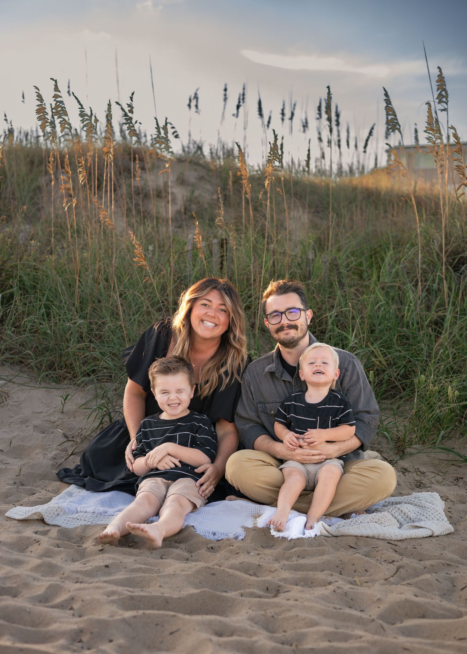 Family of four smiling on sandy beach with sea oats, two parents holding young children at golden hour.