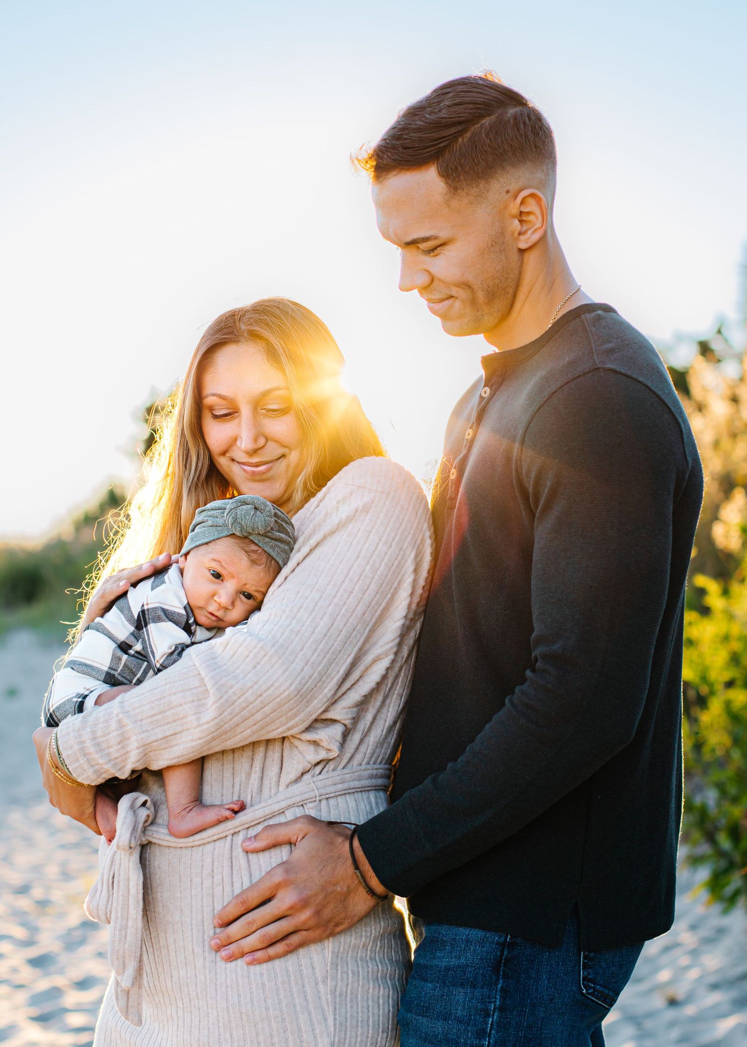 A young couple smiles at their newborn baby on a sunny beach, bathed in warm golden light.