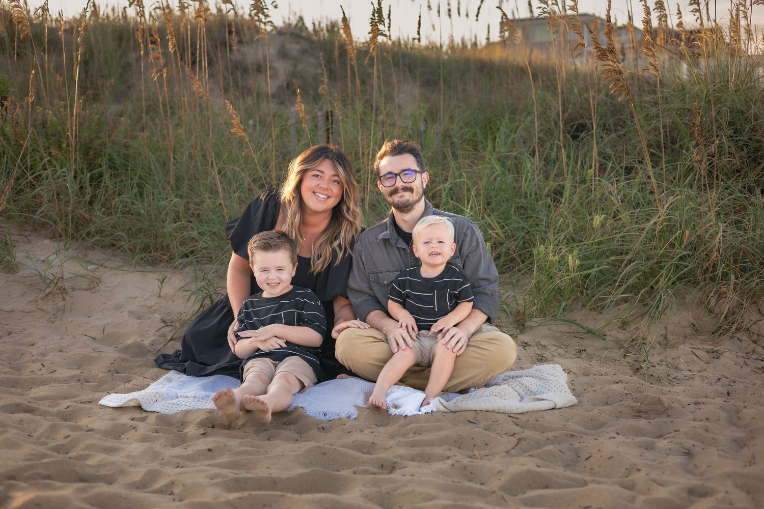 Family of four smiling on sandy beach blanket surrounded by dune grass during golden hour sunset.