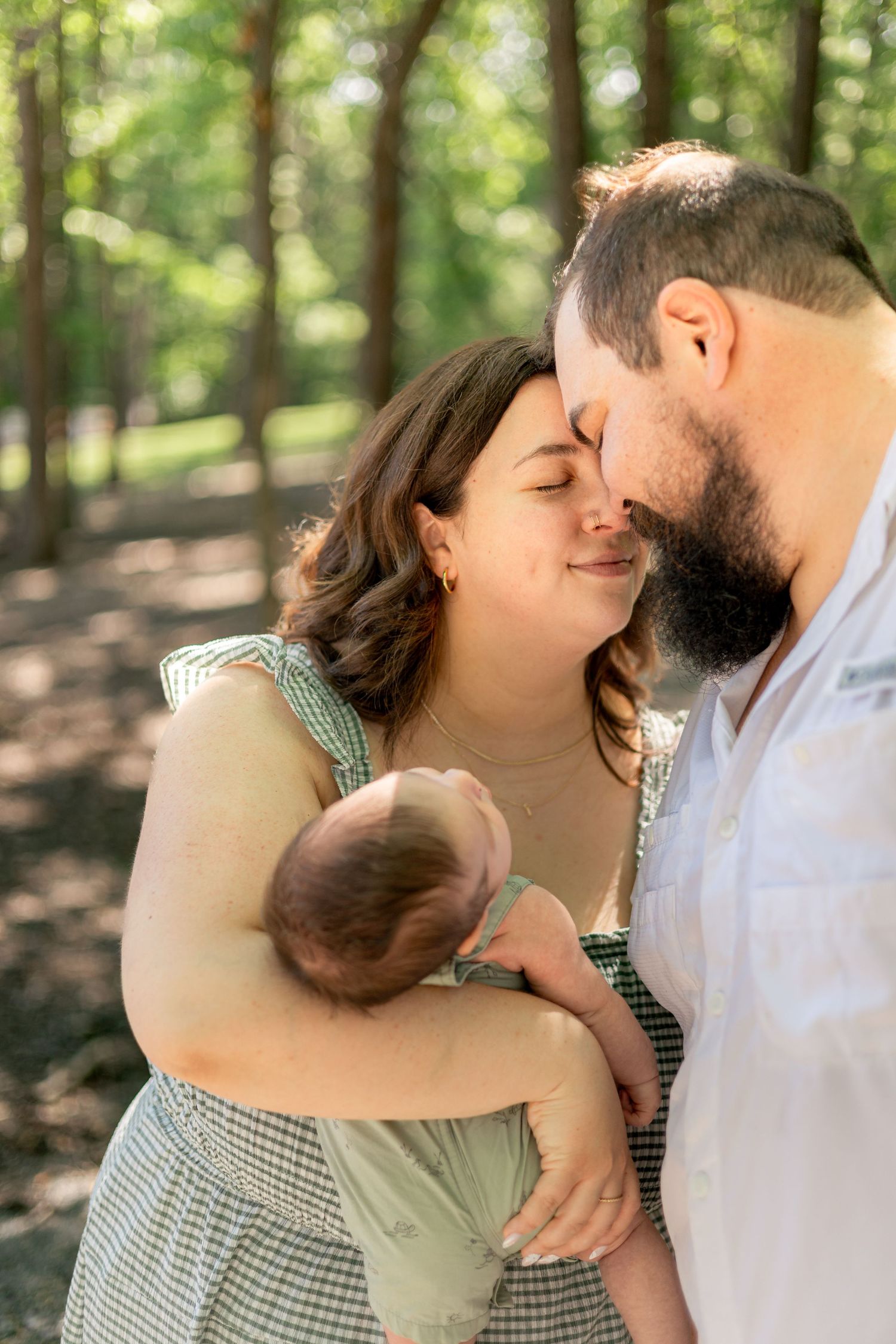 A couple sharing an intimate forehead touch while cradling their newborn baby outdoors in a sunlit forest.