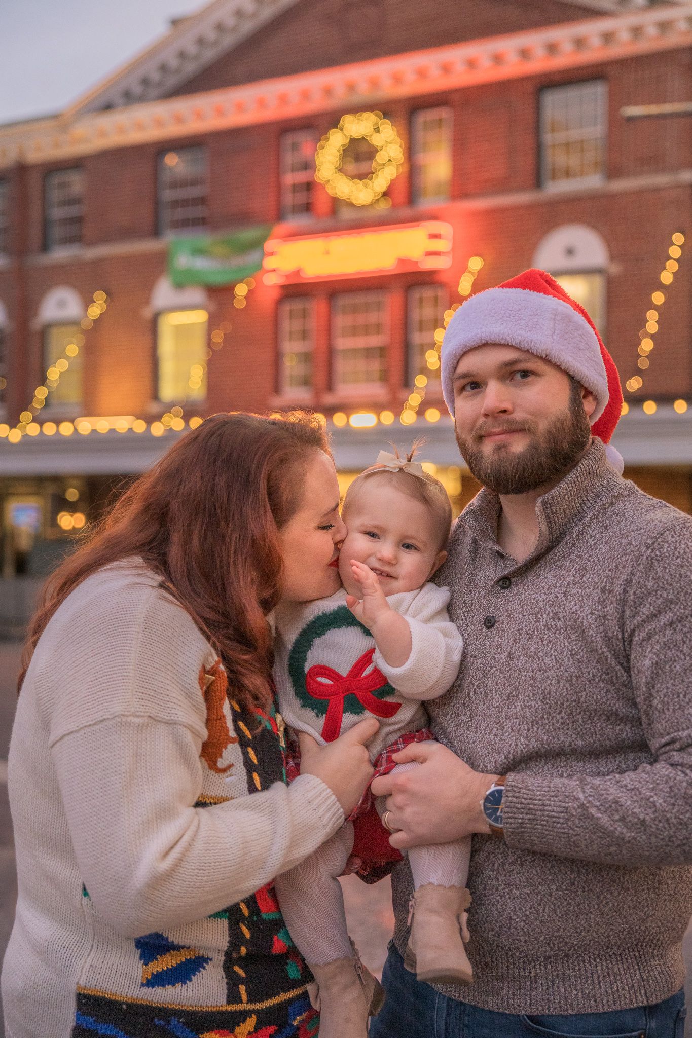 Family in Christmas sweaters holds baby outdoors near festive lit building at dusk.