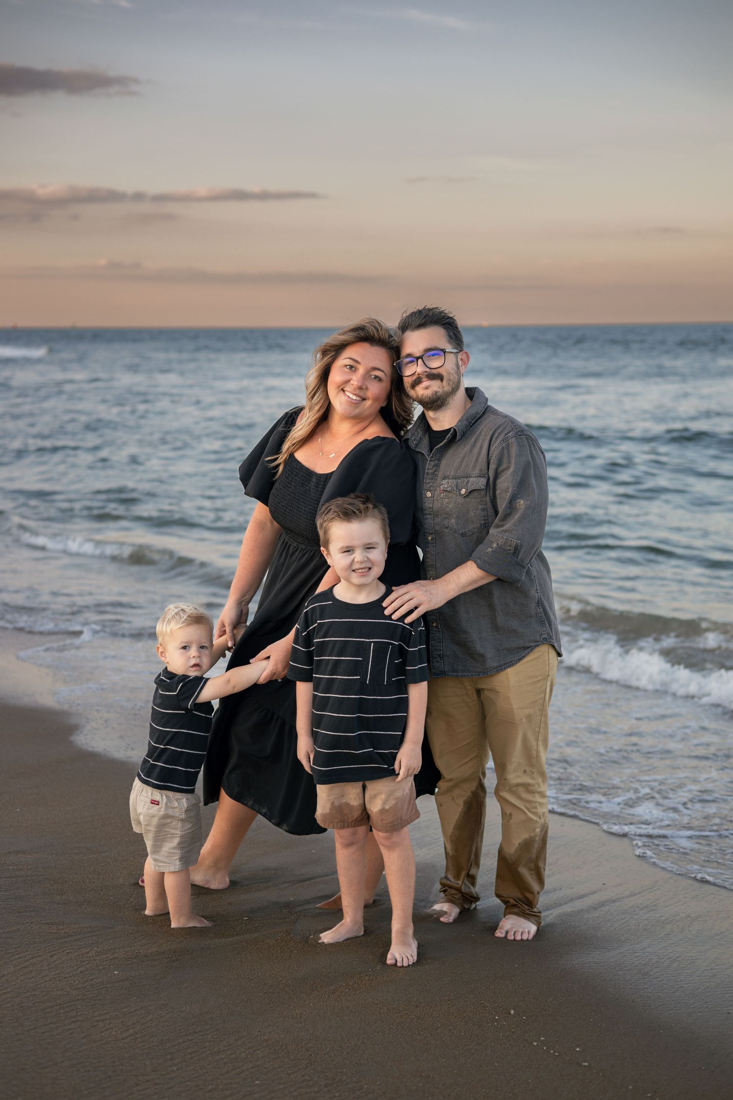 Family of four posing on beach at sunset, parents and two young boys in matching striped shirts, ocean waves behind them.
