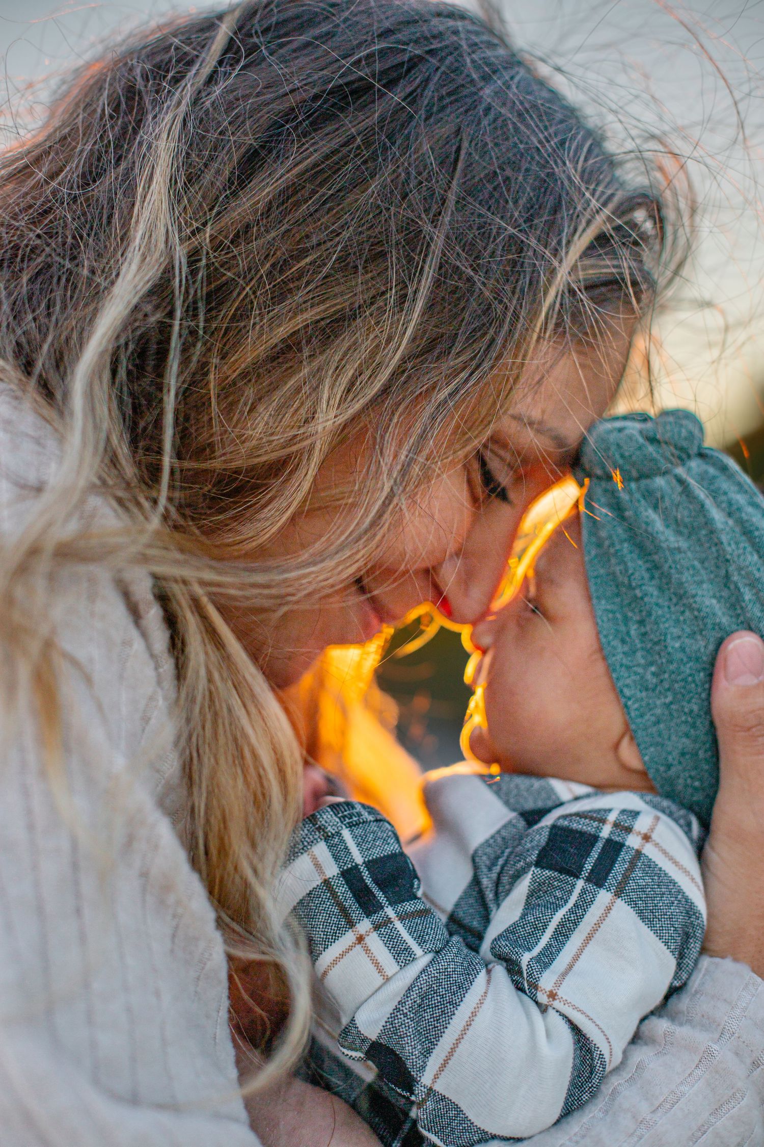 A mother tenderly touches foreheads with her baby wearing a teal hat, bathed in warm golden sunset light.