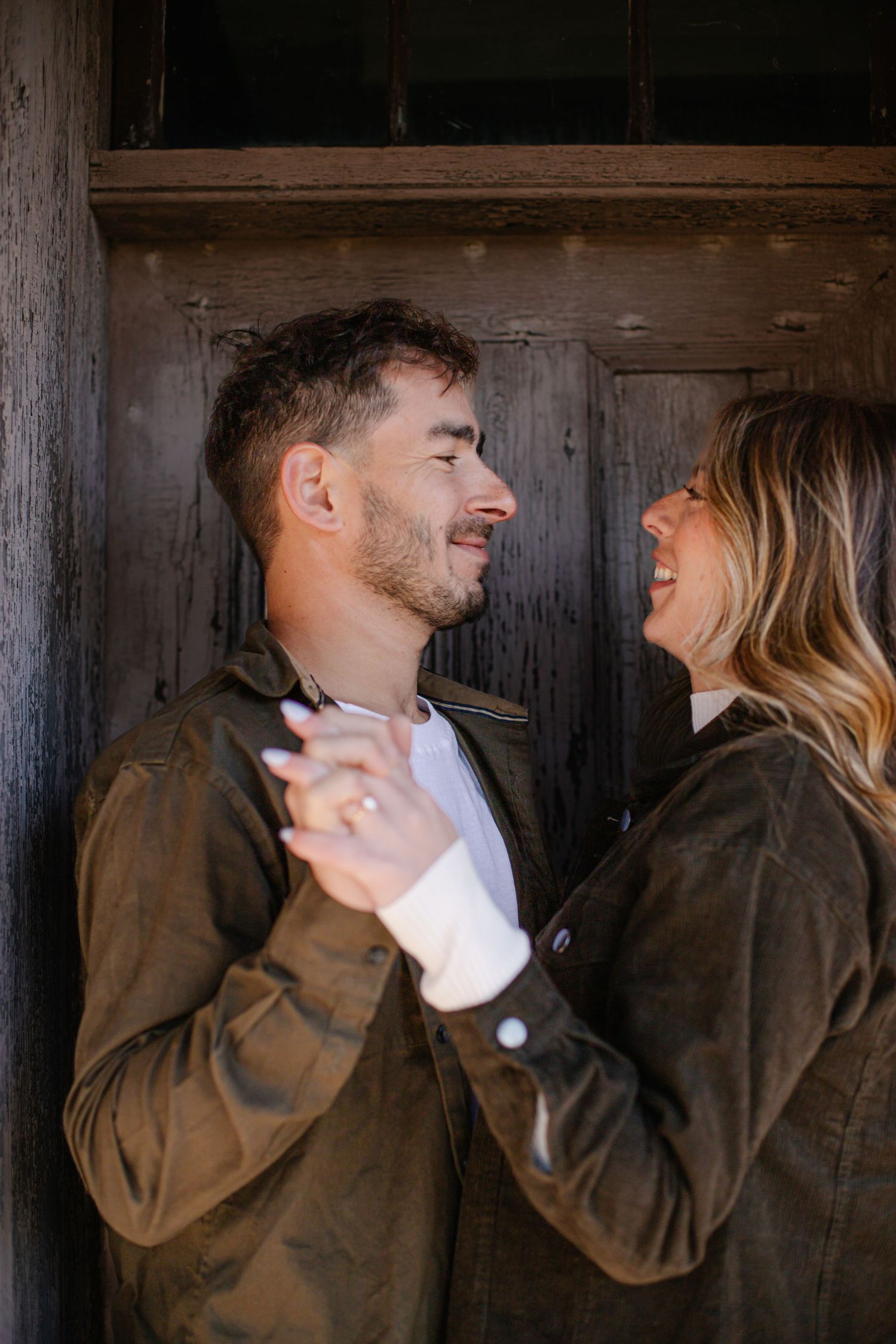 Happy couple holding hands and smiling at each other in matching brown jackets against a rustic wooden door.