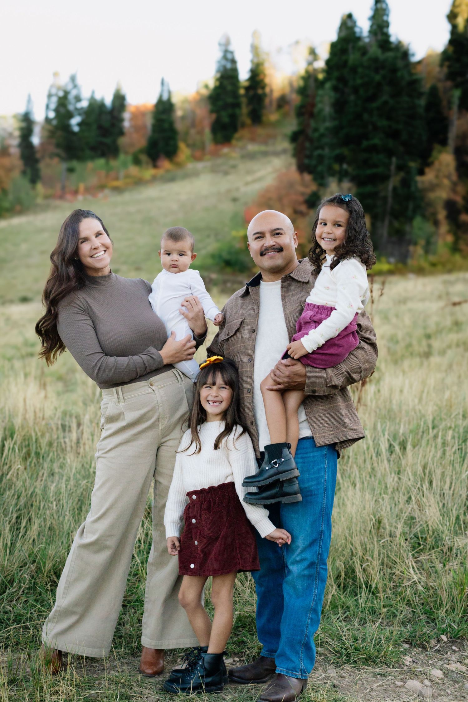 Happy family of five posing outdoors in a meadow with autumn trees, parents holding three young children.