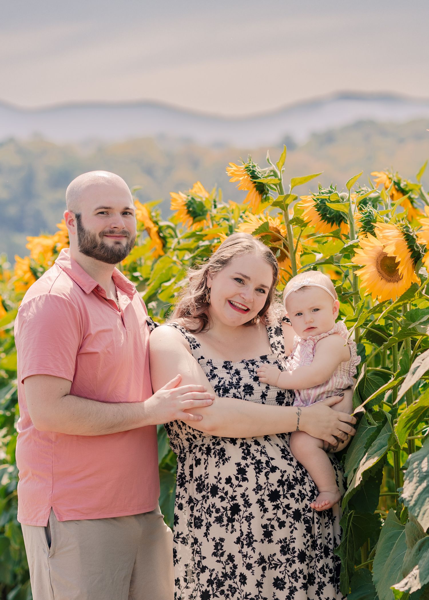 Family portrait in sunflower field with parents holding baby girl, mountains in background.